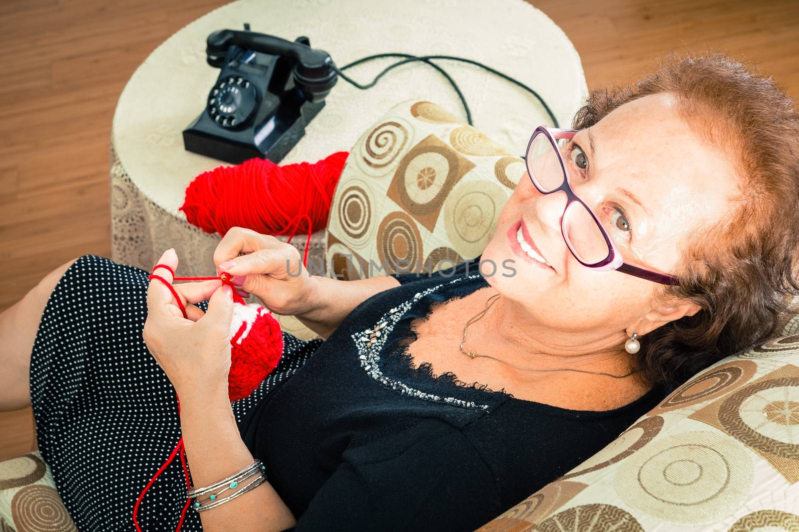 Senior Grandmother sitting in her favourite armchair enjoying her knitting as she waits for a phone call looking up to smile at the camera, viewed from above