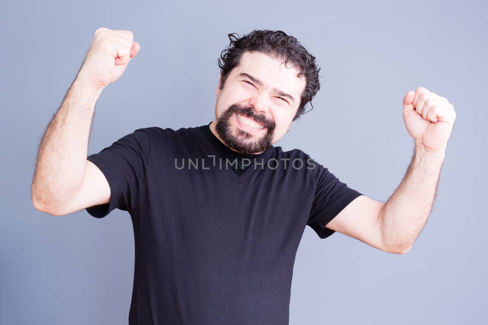 Single handsome bearded man in black shirt holding fists up with elated expression over gray background