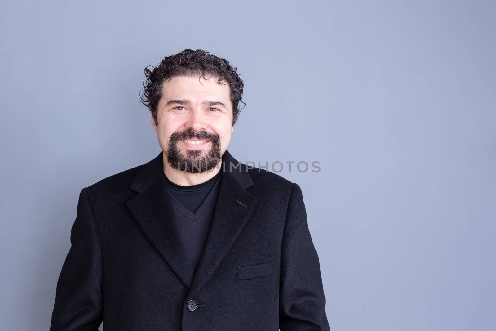 Single smiling handsome dark haired and bearded middle aged man wearing black shirt and blazer over gray background with copy space