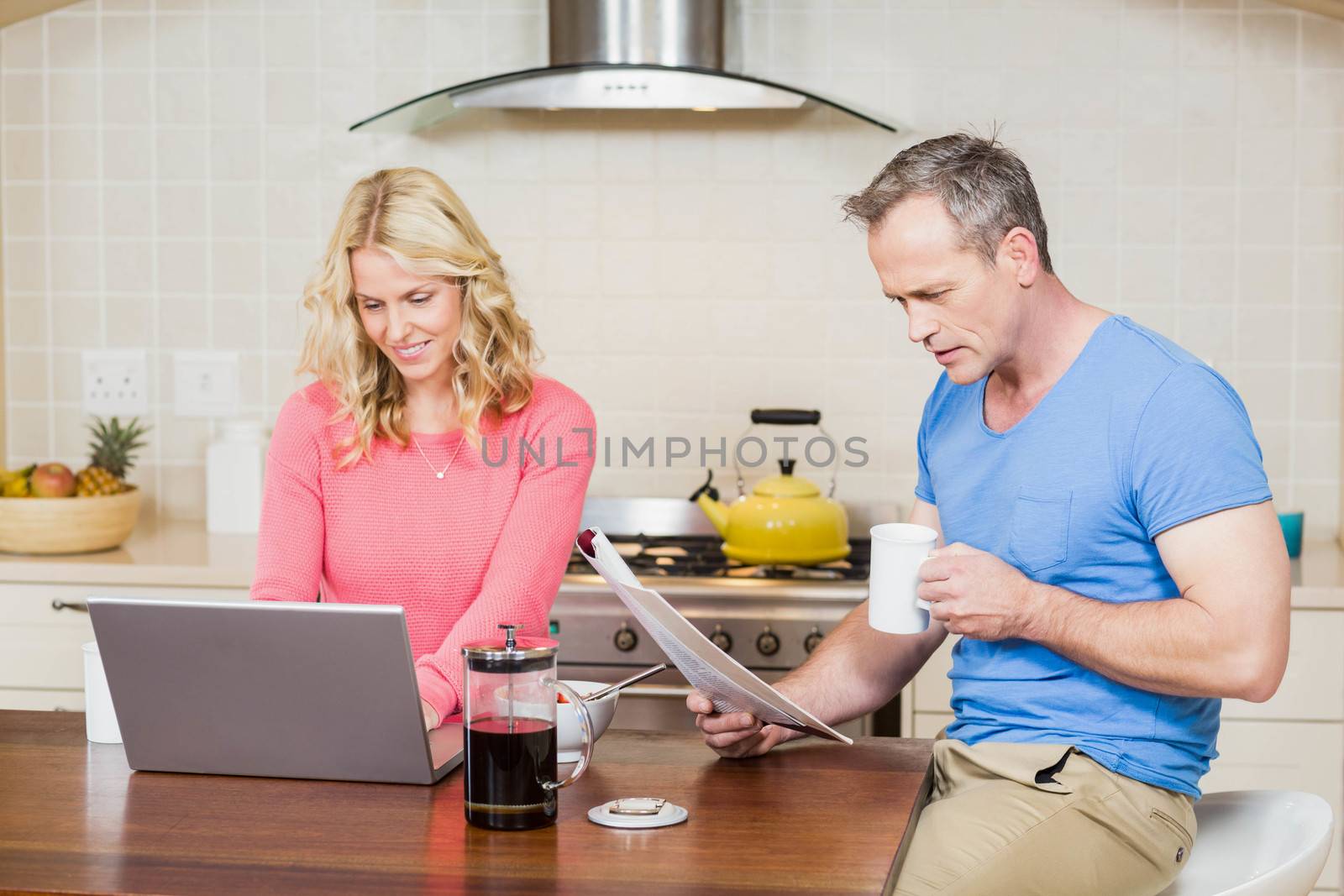 Mature couple eating breakfast while using laptop and reading in the kitchen