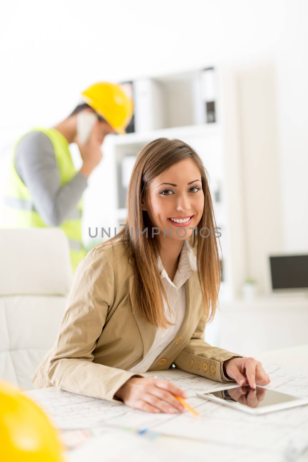 Smiling female architect with blueprint and tablet sitting at desk in office. Looking at camera.