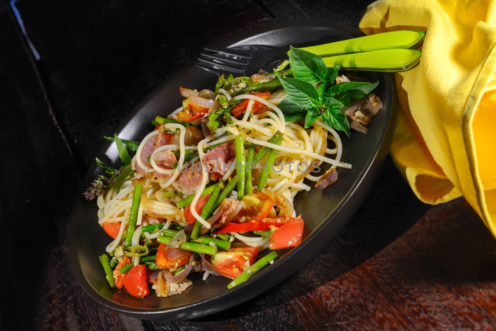 Spaghetti with asparagus tomatoes bacon and herbs on wood table in a black plate