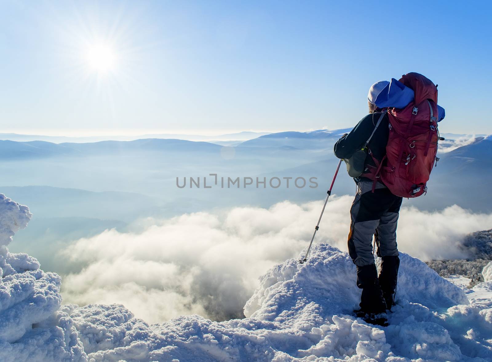 A Tourist with a backpack standing on a mountain top above the clouds and looking at the view. Direct sunlight. Clear blue sky. Winter. Mountaineering. Ukraine. Carpathians