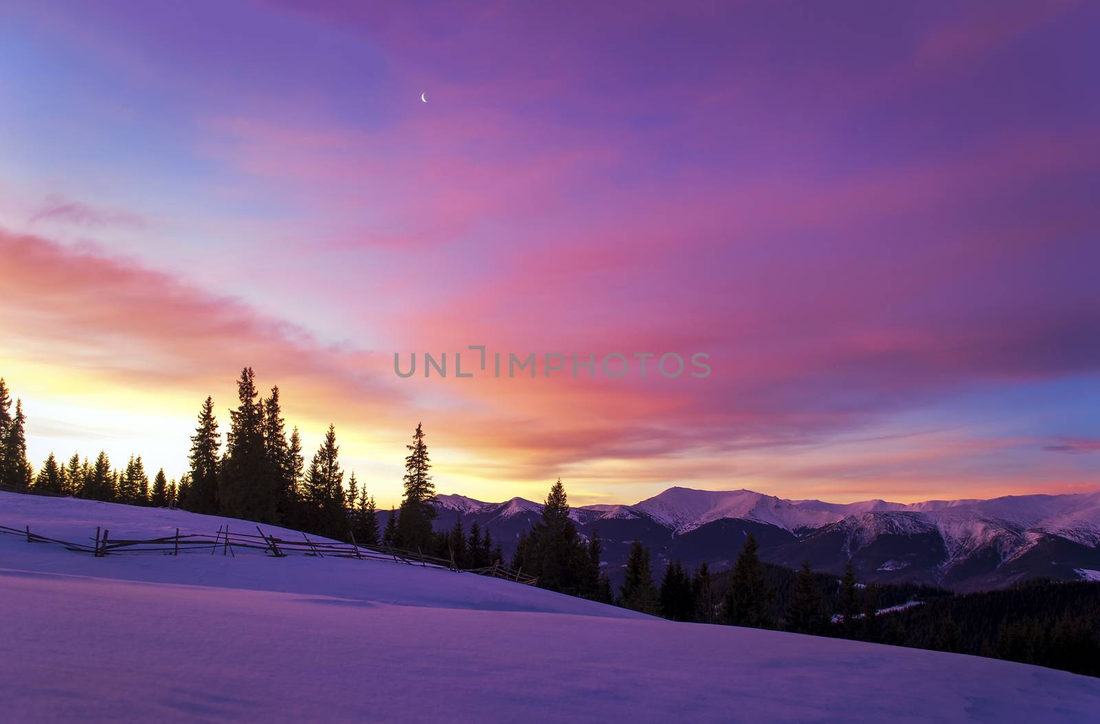 Winter morning landscape. The sun rises behind trees and lights up the clouds. Mountain ridge. The moon In the sky. Wooden fence in snowy field. Twilight. Purple colors. Romantic mood