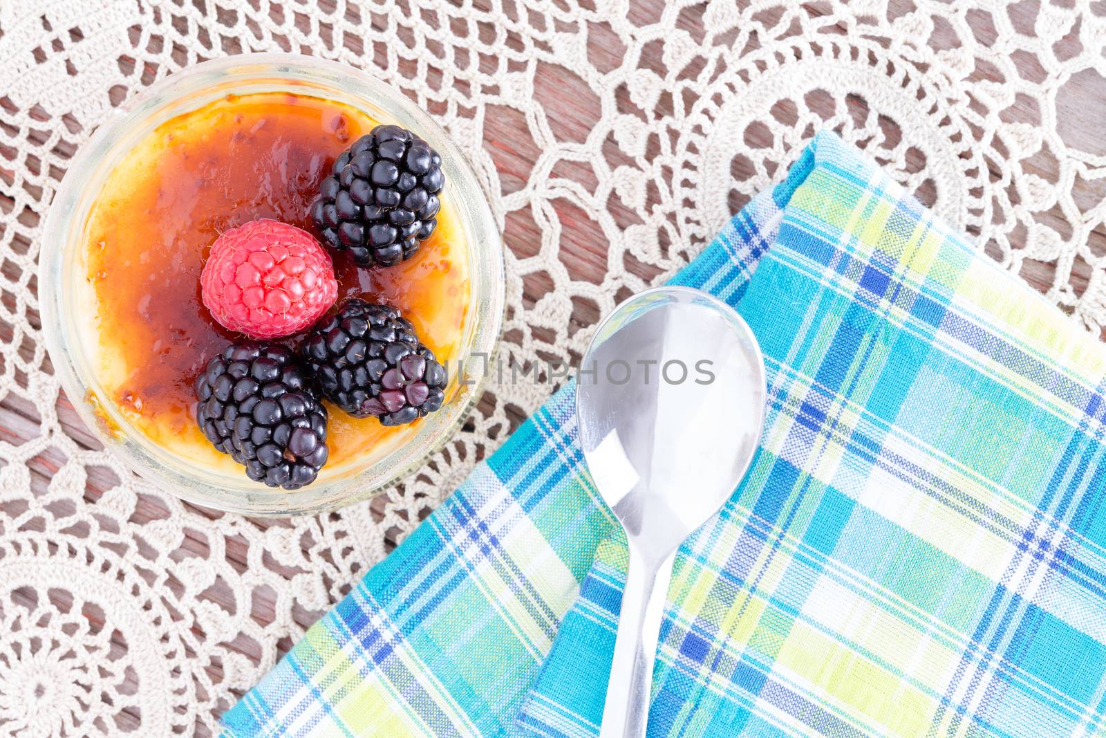 Delicious caramelized custard topped with red and black raspberries from point of view angle with spoon over doily and blue napkin