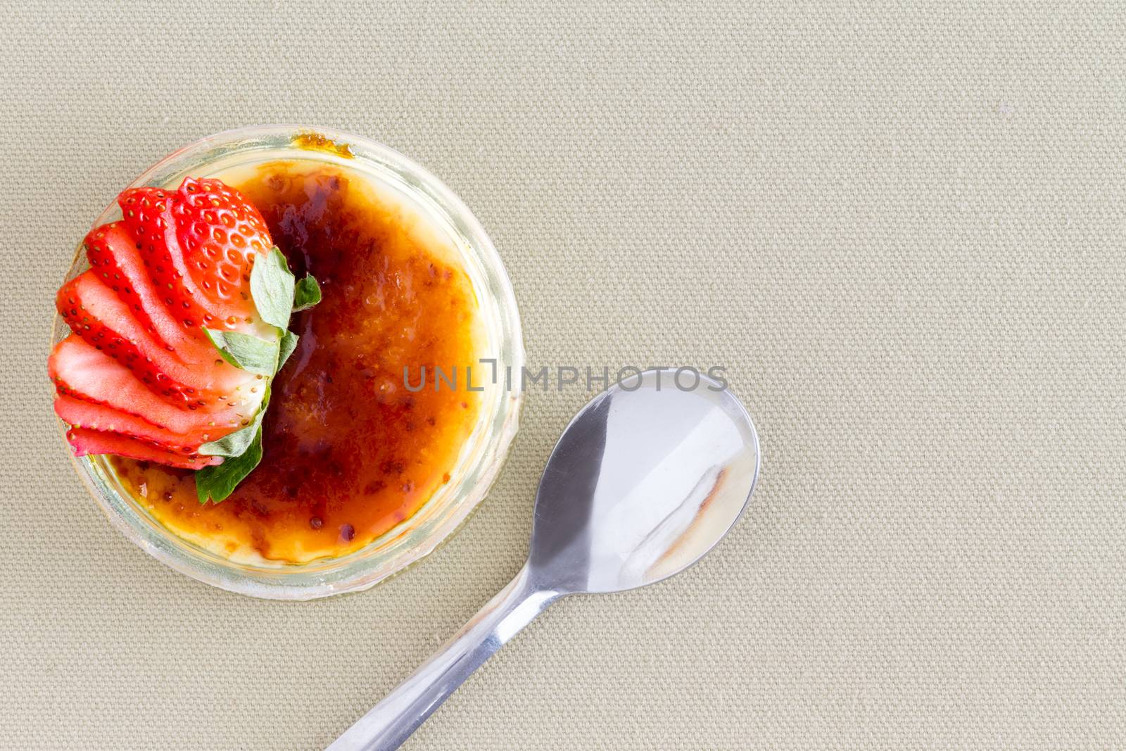 Delicious custard dessert topped with strawberry slices from point of view angle with spoon over brown tablecloth