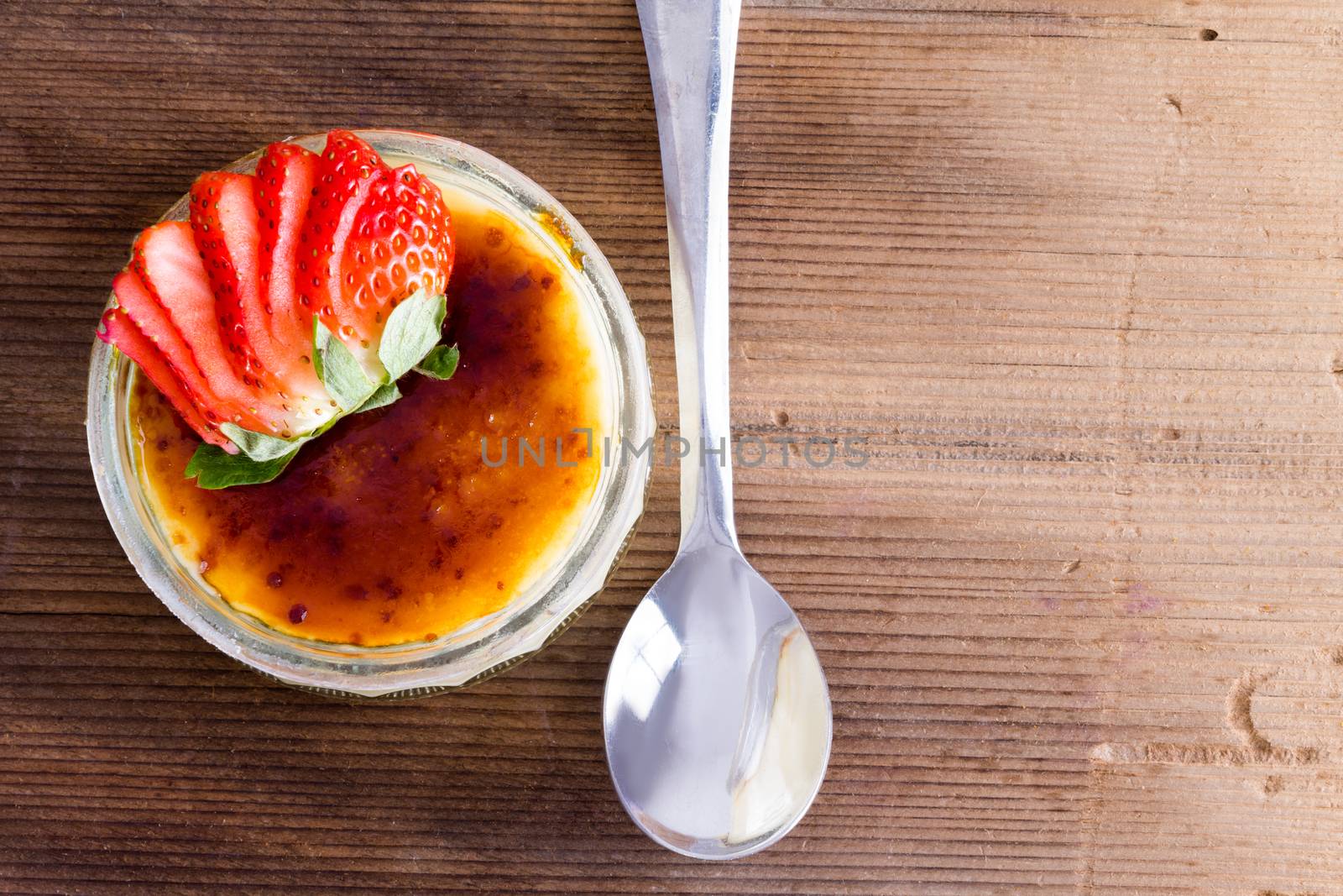 Appetizing custard dessert topped with sliced strawberry from above angle with spoon over wood background