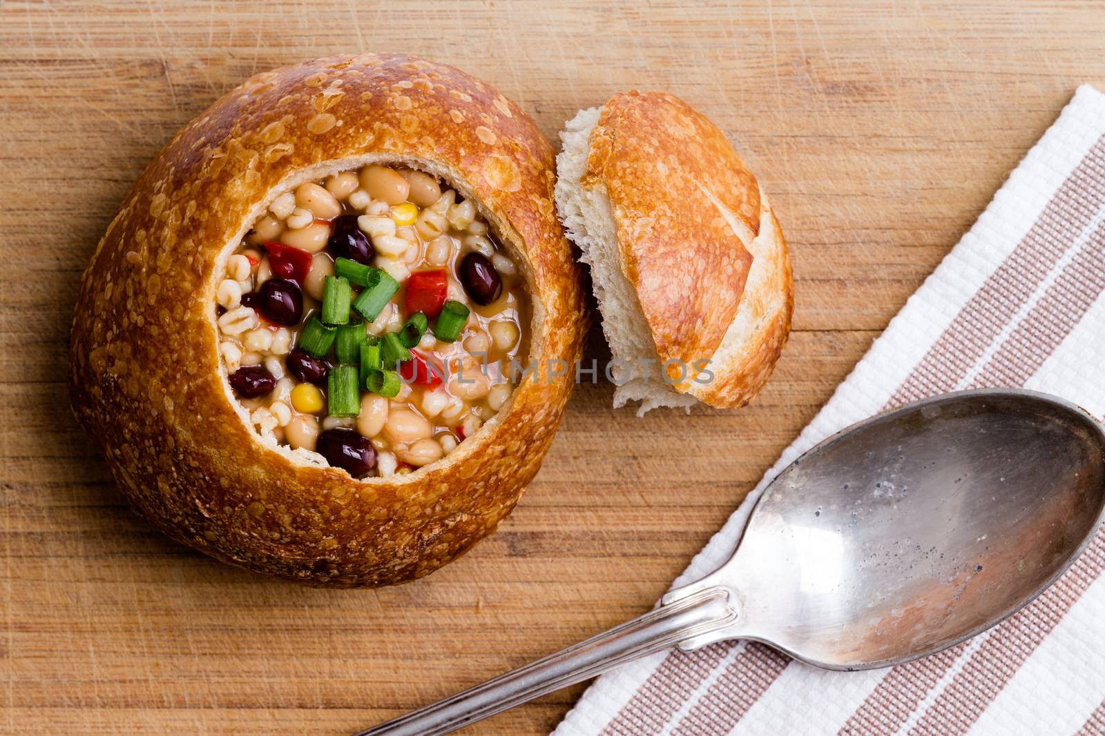 Top view of delicious bread bowl with beans, barley, corn and vegetables next to lid and spoon on table