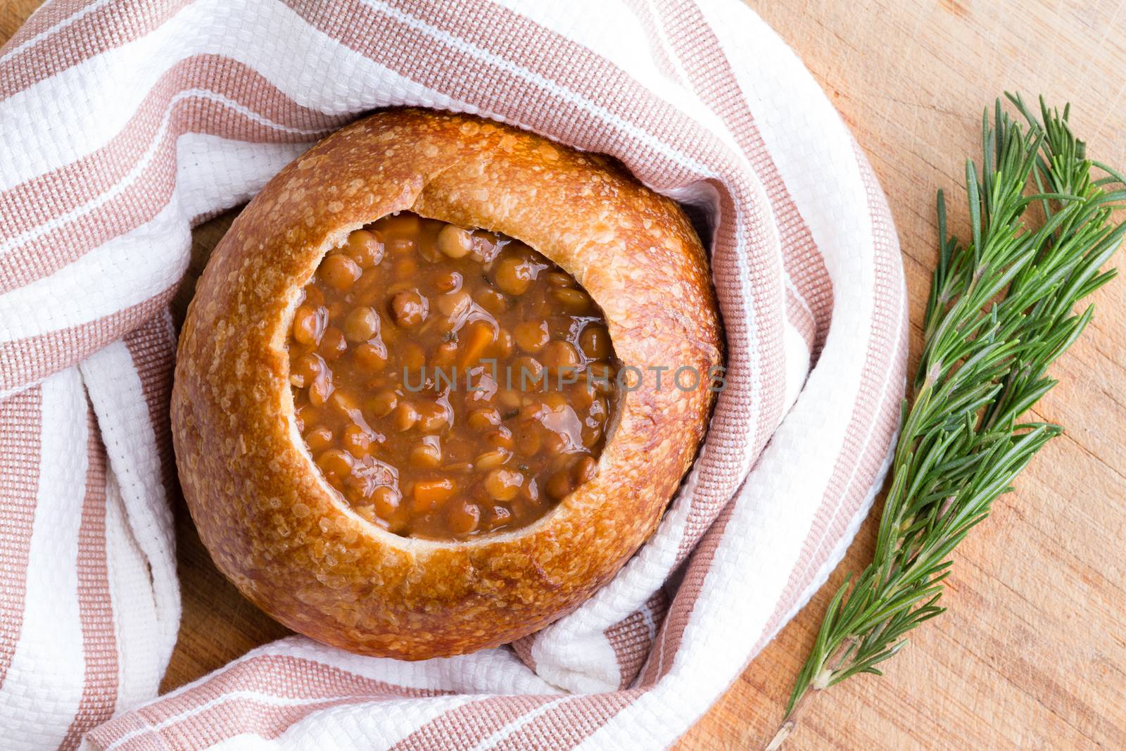 Freshly cooked warm bread bowl full of lentil soup wrapped in cloth napkin next to sprigs of rosemary on table