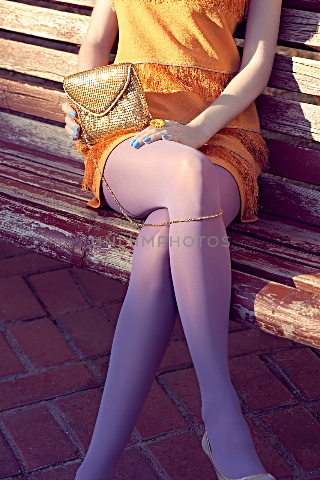 Fashion urban beauty people, woman, outdoor. Playful glamor hipster girl on bench in pantyhose, stylish orange dress with gold clutch, sunny day. Creative unusual pose, lifestyle. Vivid disco party 