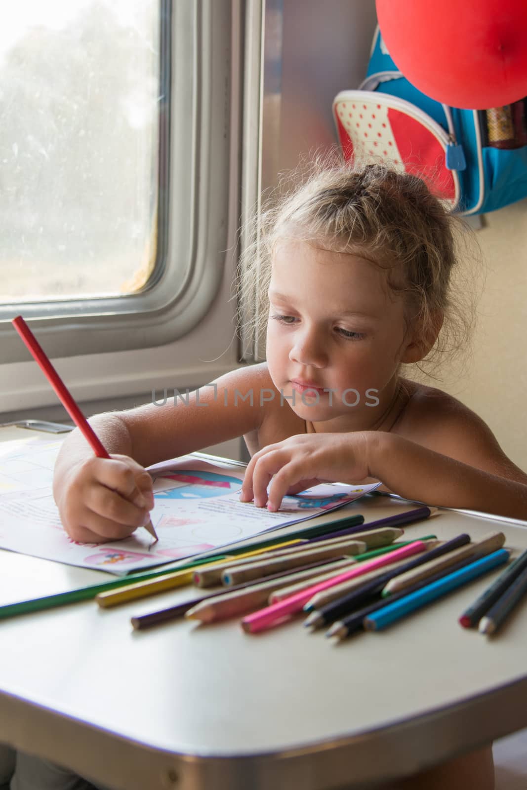 Four-year girl draws pencils in a magazine at the table while in the second-class train carriage