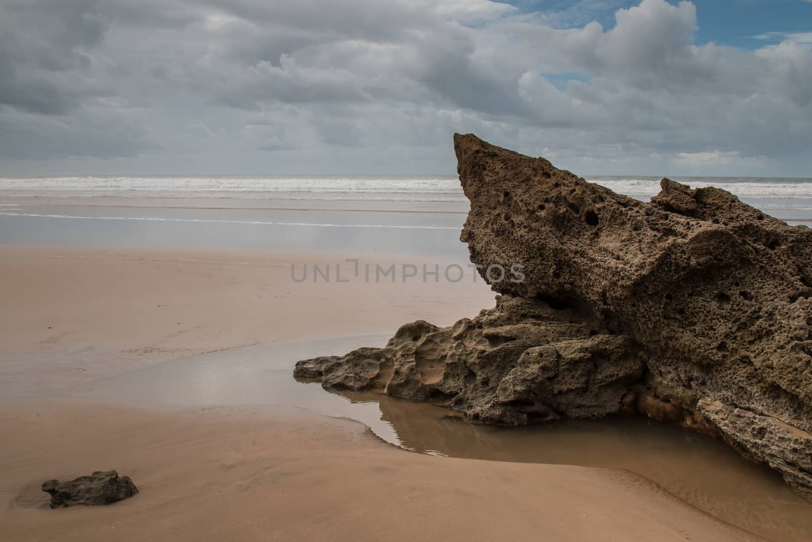 Rocks on the sandy beach in the foreground. Windy autumn day with many clouds. Waters of Atlantic Ocean in the background. Moulay Bouzerktoun, Morocco.