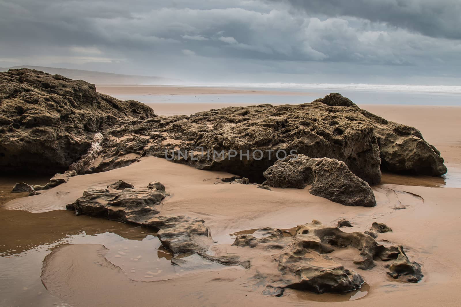 Rocks on the sandy beach in the foreground. Windy autumn day with many clouds. Waters of Atlantic Ocean in the background. Moulay Bouzerktoun, Morocco.