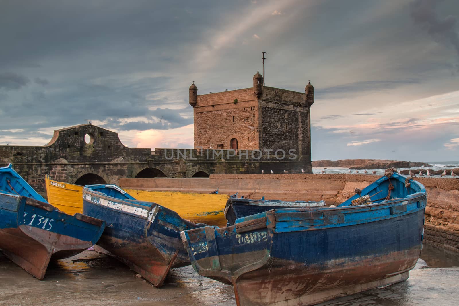 Parked boats in the port of Essaouira. Tower as a part of city fortification in the background. Early morning cloudy sky.