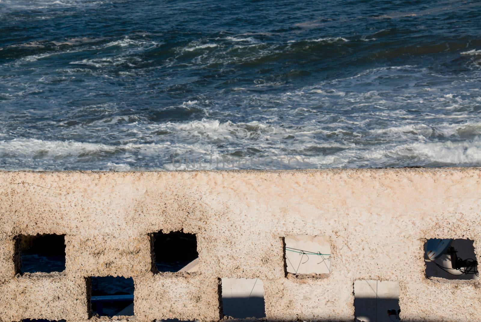 Fence enlightened by the sun and wild water of the ocean in the background. View from a roof of a building.