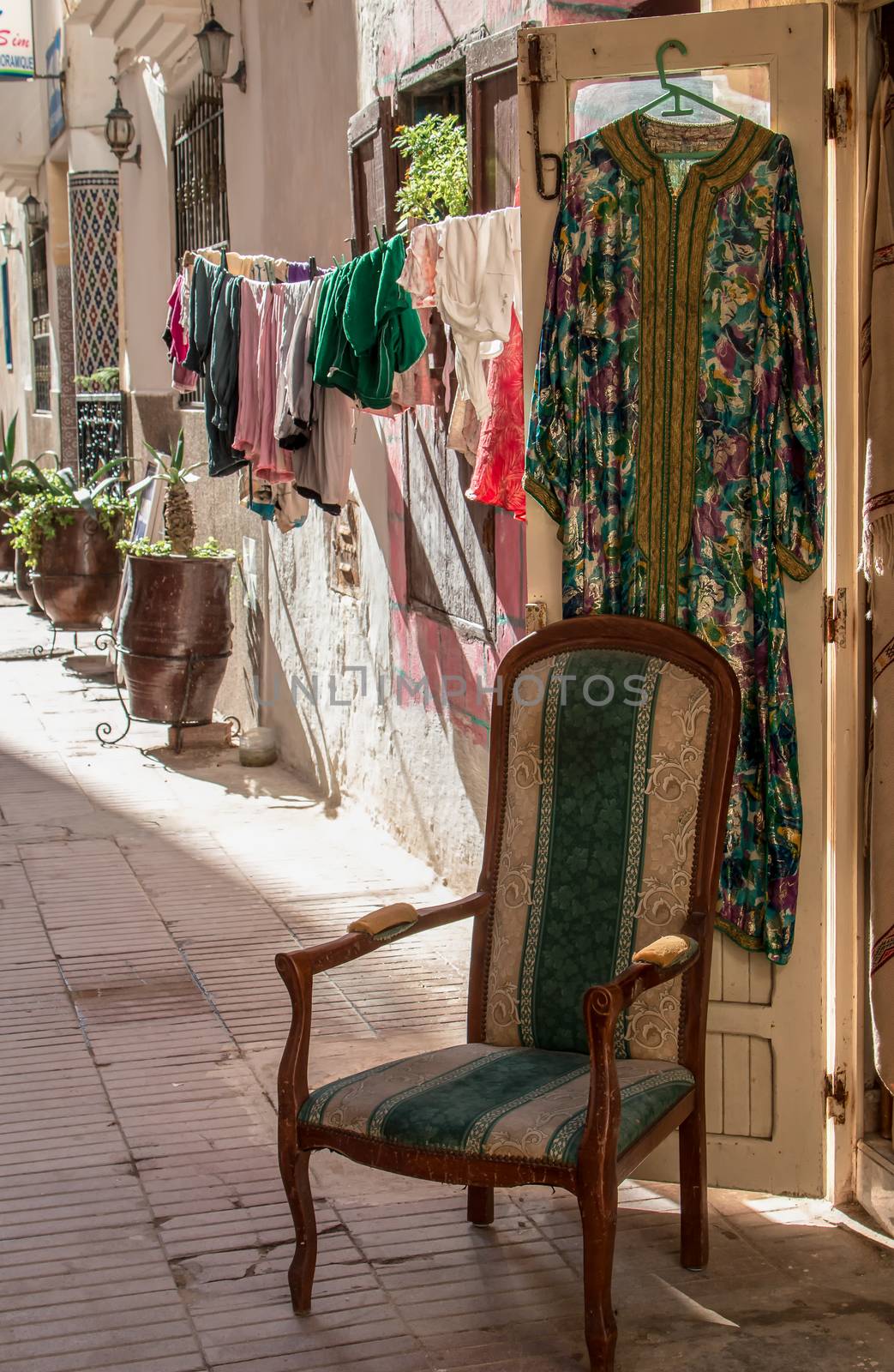 Street with a shop, where are dresses hanging on the door and there is a comfortable old style chair. Behind laundry hanging to dry. Essaouira, Morocco.