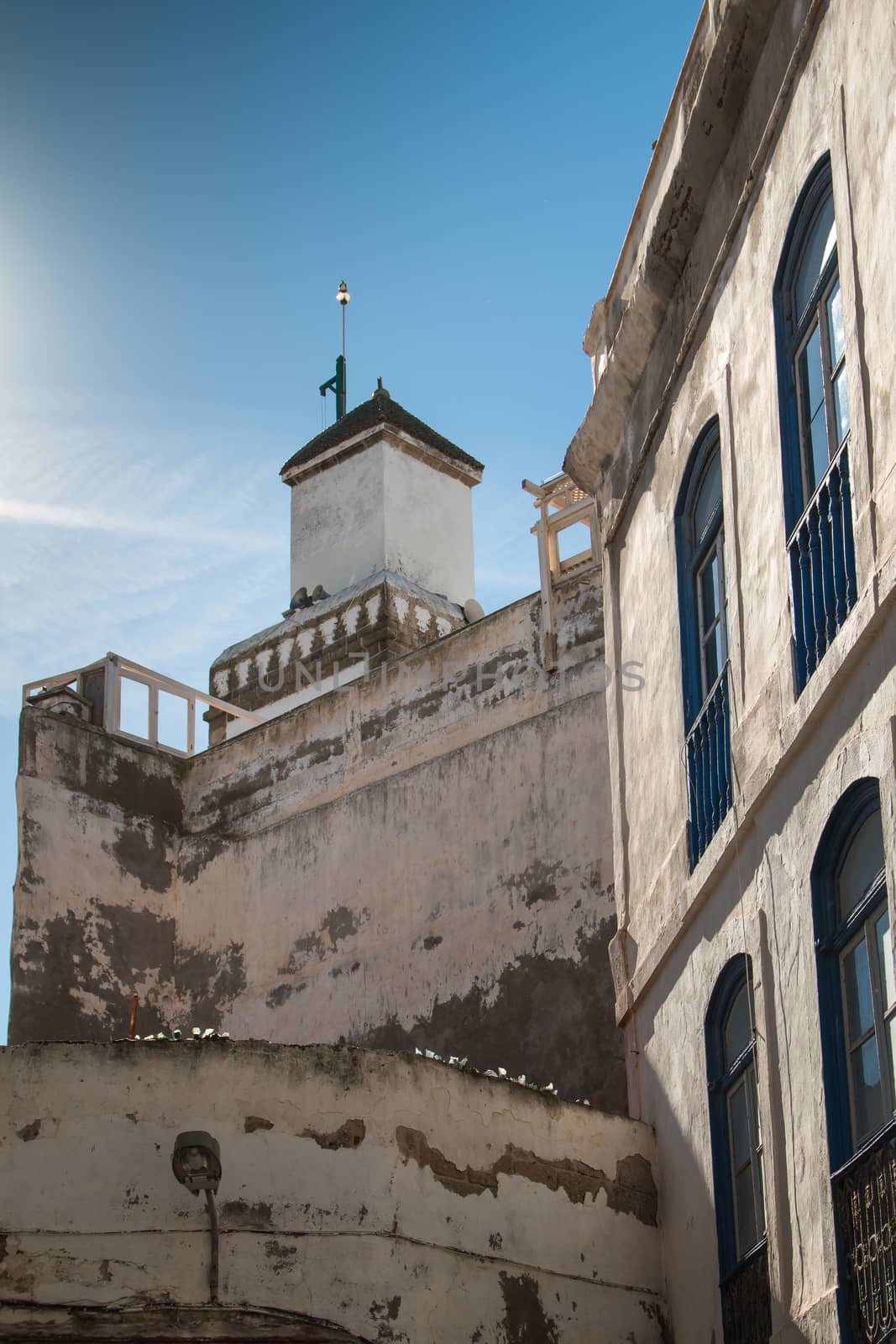 Street with high buildings with blue framed windows. In the background tower-minaret of a mosque. Bright blue morning sky. Essaouira, Morocco.