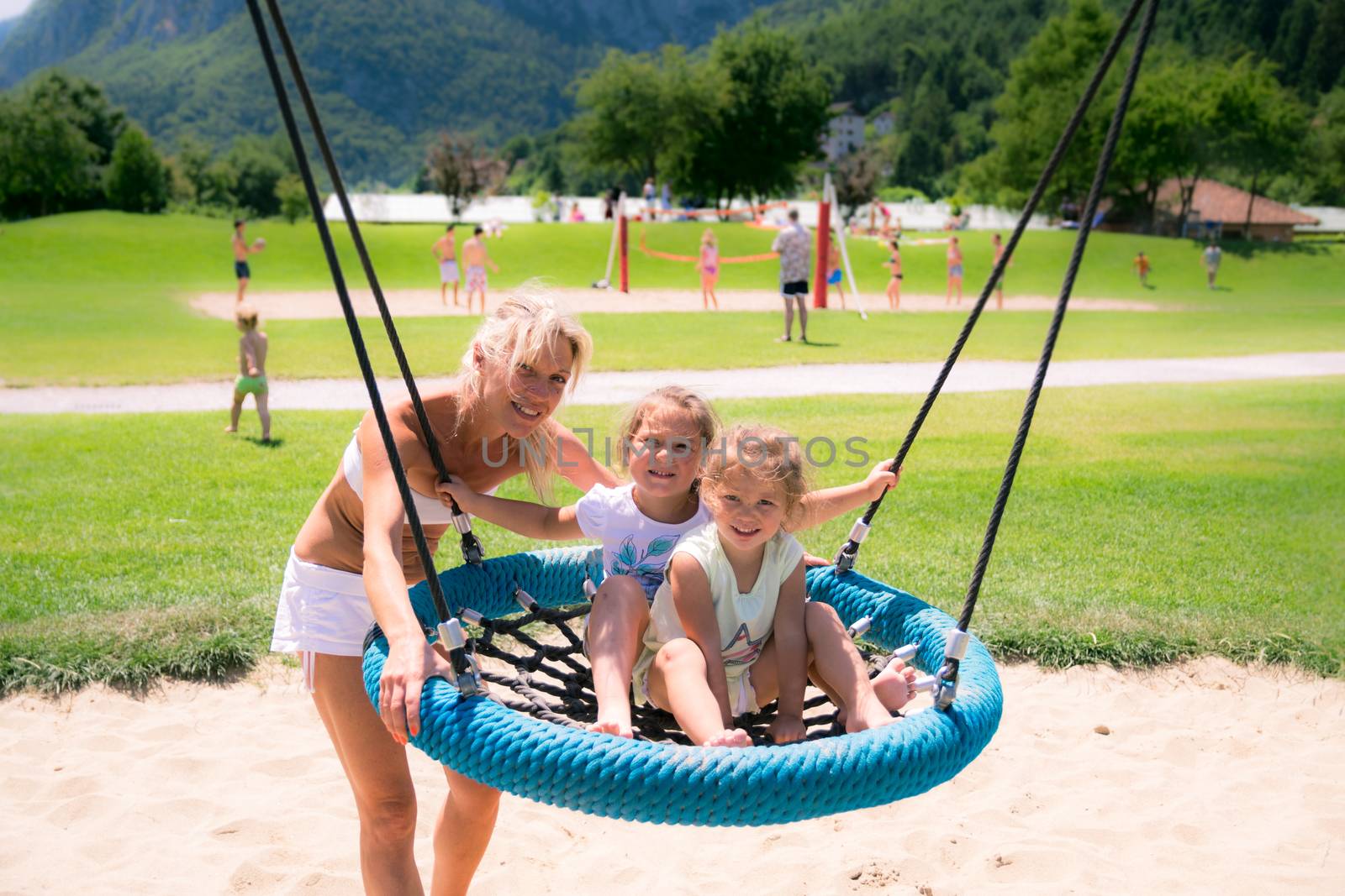 Blonde mom pushes the girls on the round rope swing in a playground.