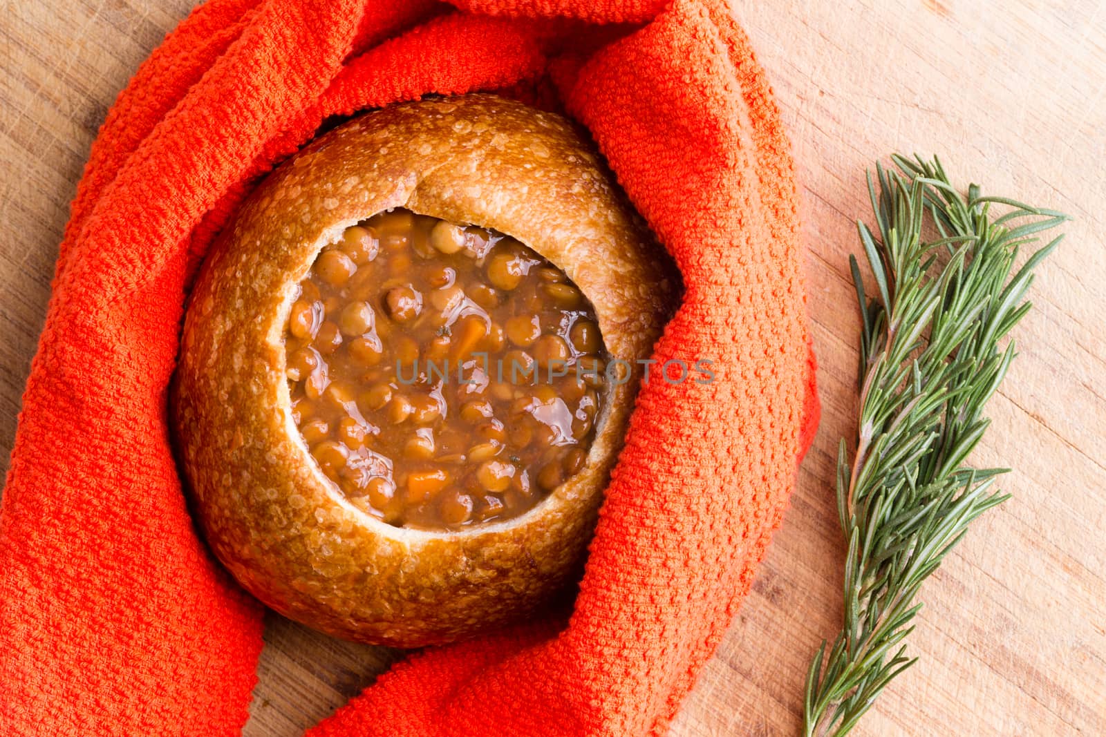 Freshly cooked warm bread bowl full of lentil soup wrapped in red cloth napkin next to sprigs of rosemary on table