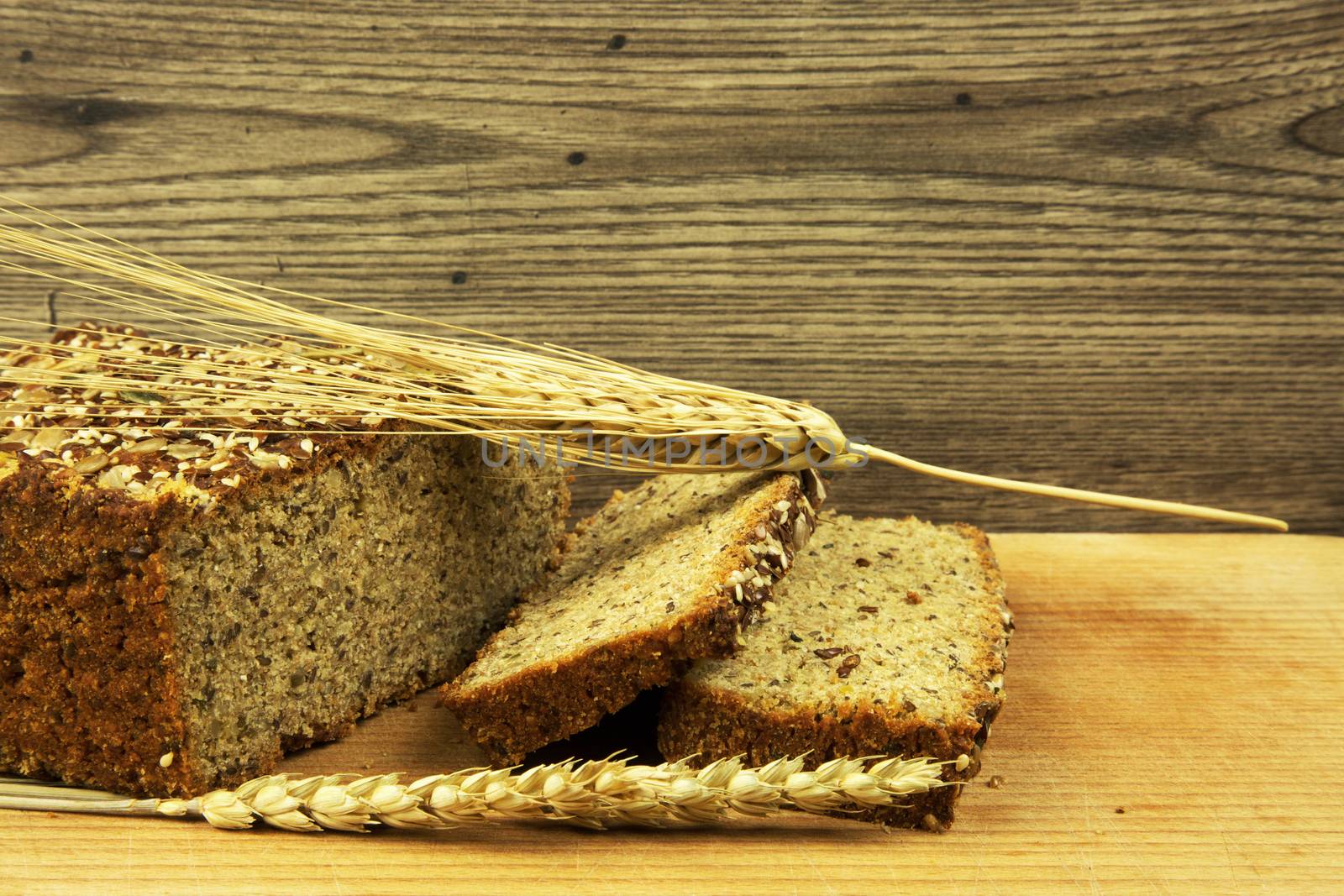 Loaf and two slices of bread with grains and wheat spikes on old wooden table. Close, horizontal view.