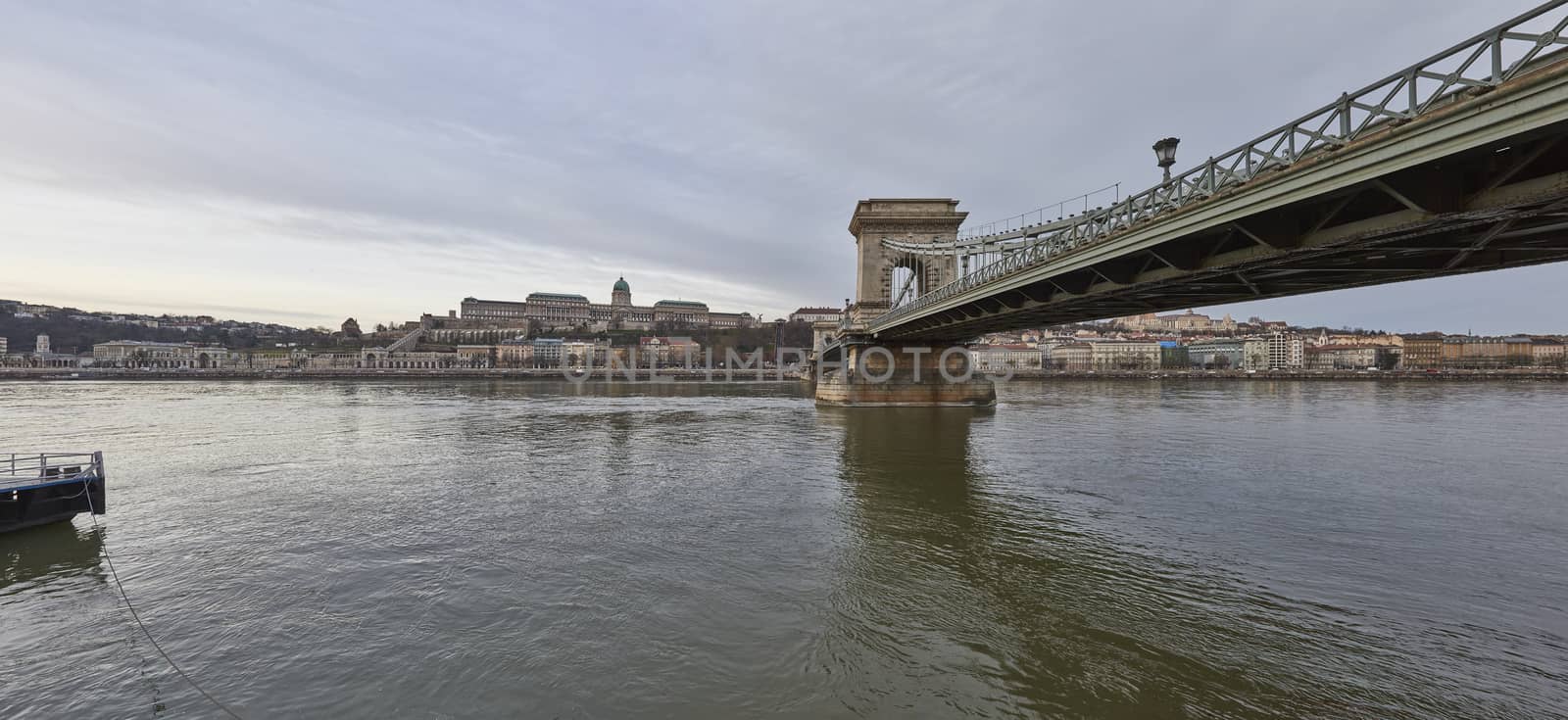 Composite panoramic low angle shot of Szechenyi Chain Bridge with riverbed rocks in the foreground and Buda Castle in the background, accross Danube River. February 02, 2016 in Budapest, Hungary.