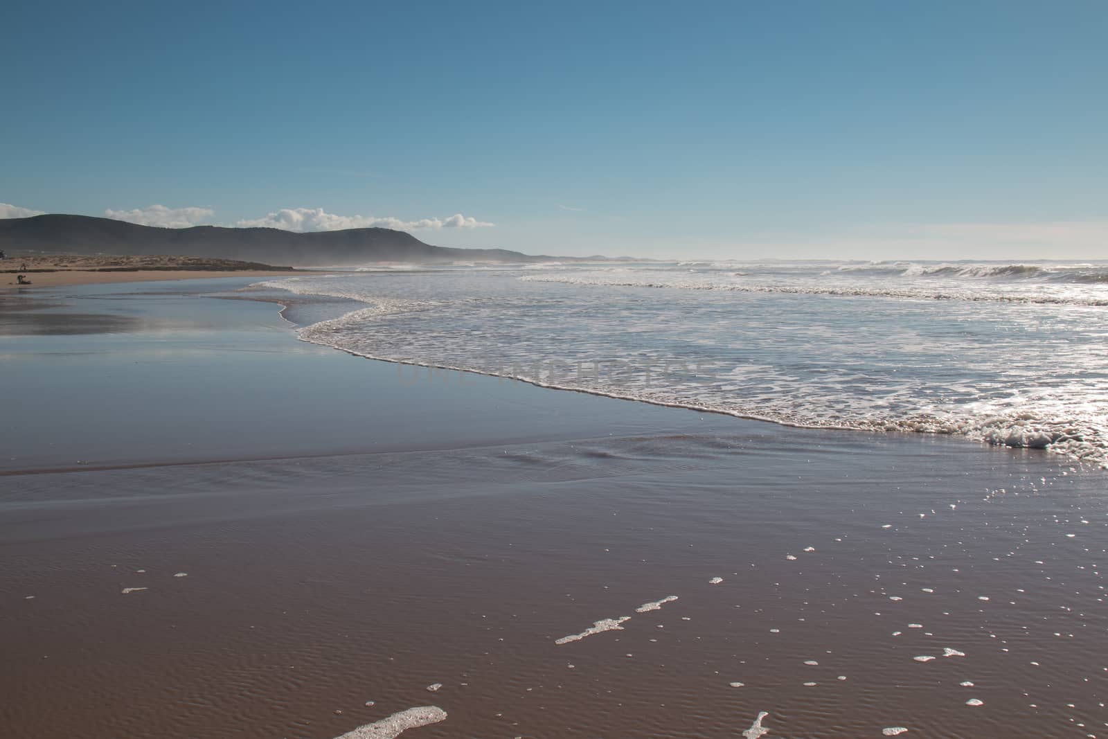 Late afternoon on the sandy beach of the Atlantic Ocean in Morocco. Waves of a tide. Mountain in the background. Blue sky with some clouds.