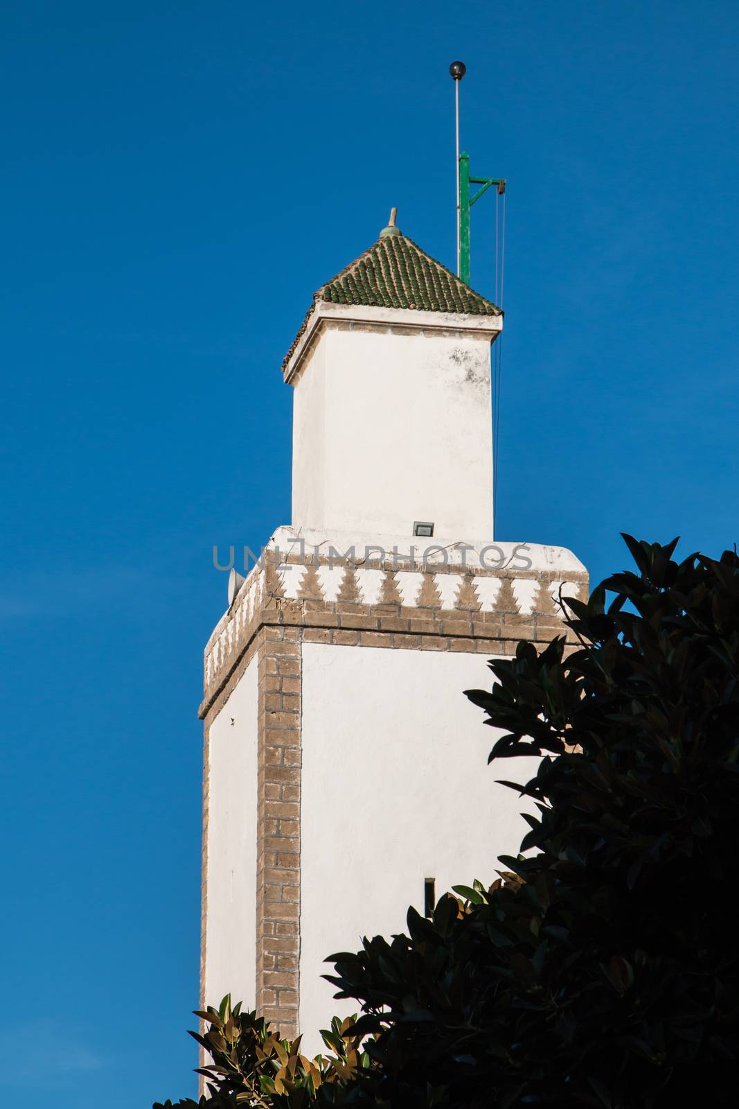 White tower of a mosque, with edges made of bricks. Green roof. Bush in the foreground. Bright blue sky. Essaouira, Morocco.