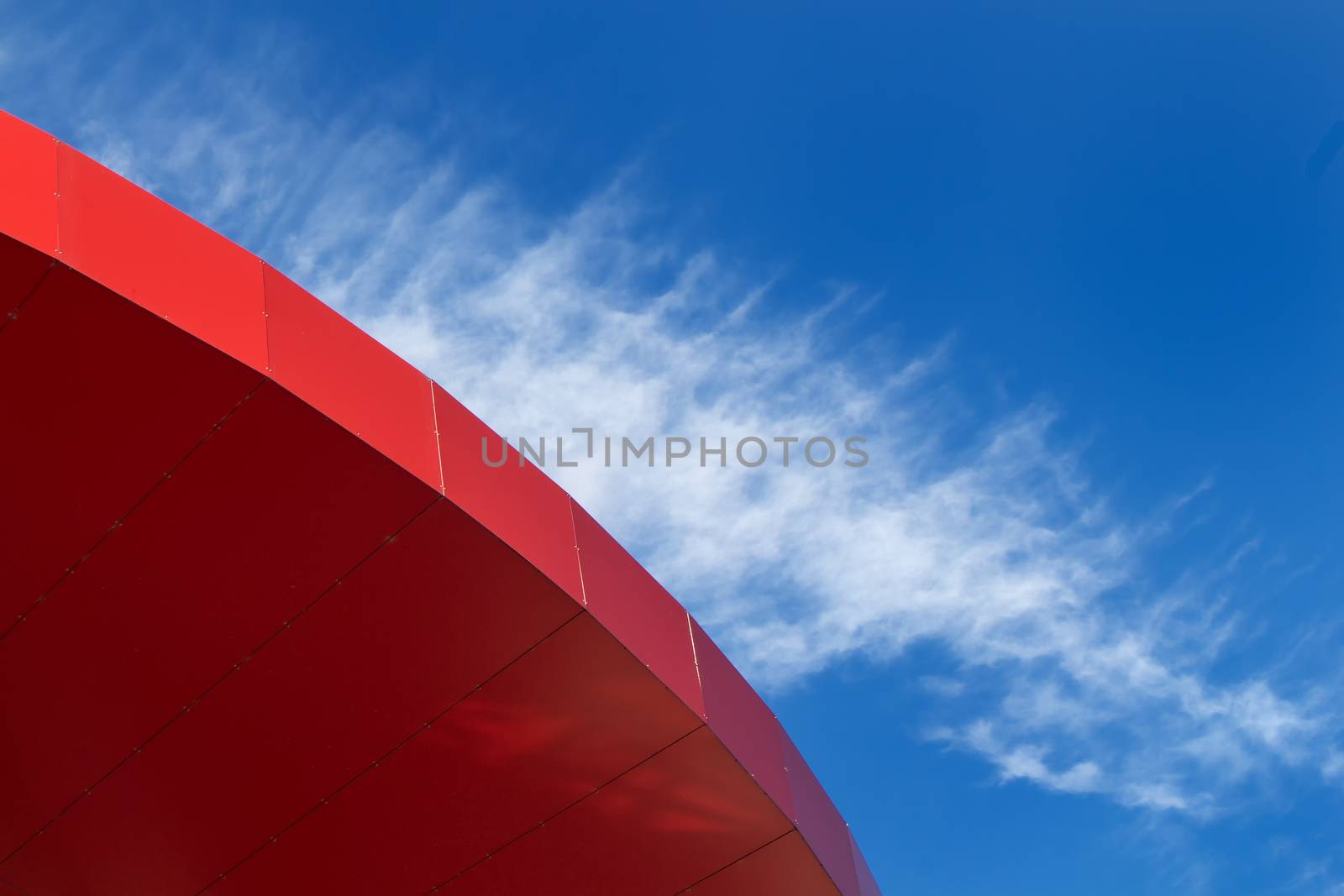 Part of a bright red modern roof with a bright blue sky in the background. Cloud lining the shape of the roof.