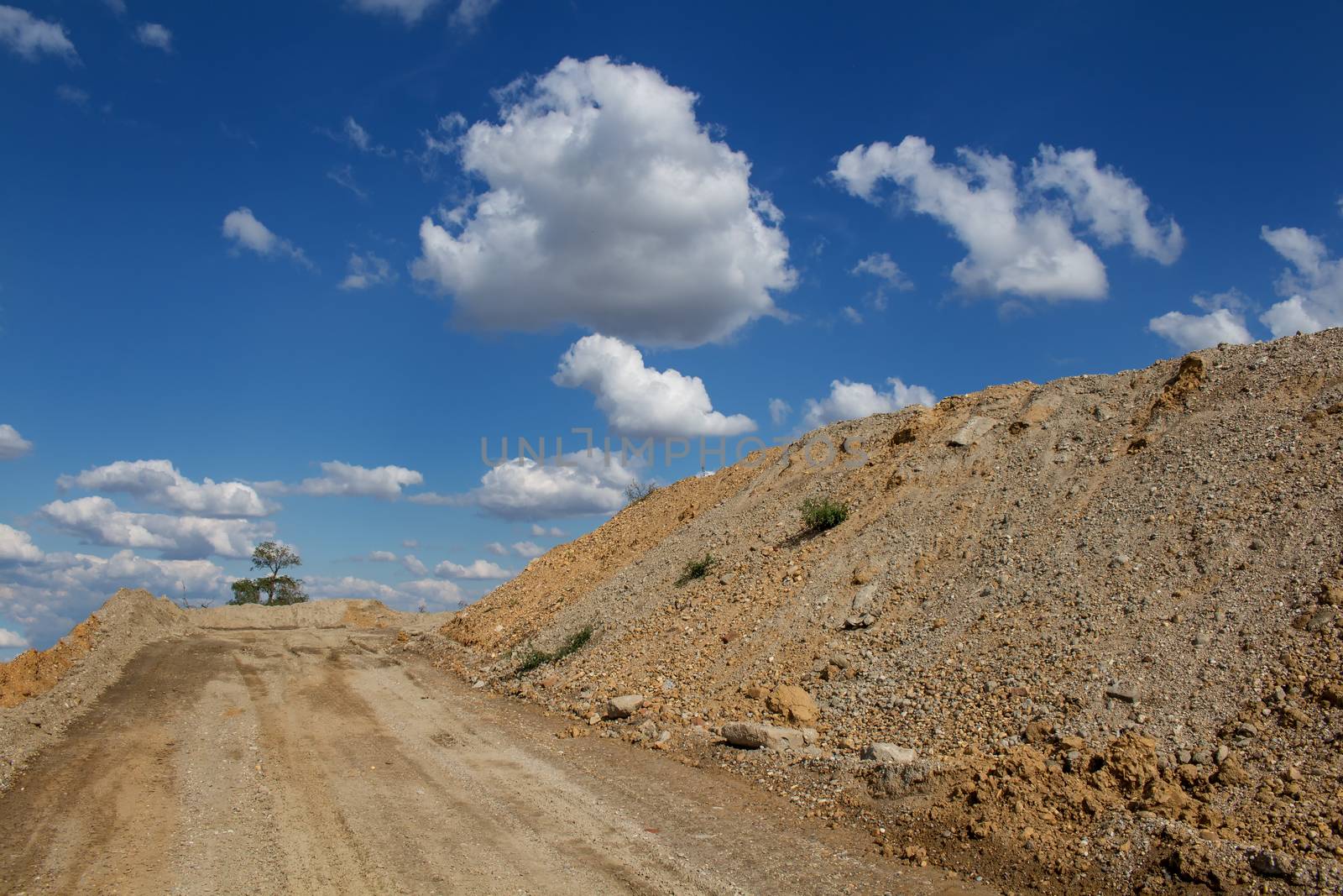 Road up the hill. Place ready for building. Devastated nature, just one tree remaining. Dry hill on the right side. Blue sky with many clouds.