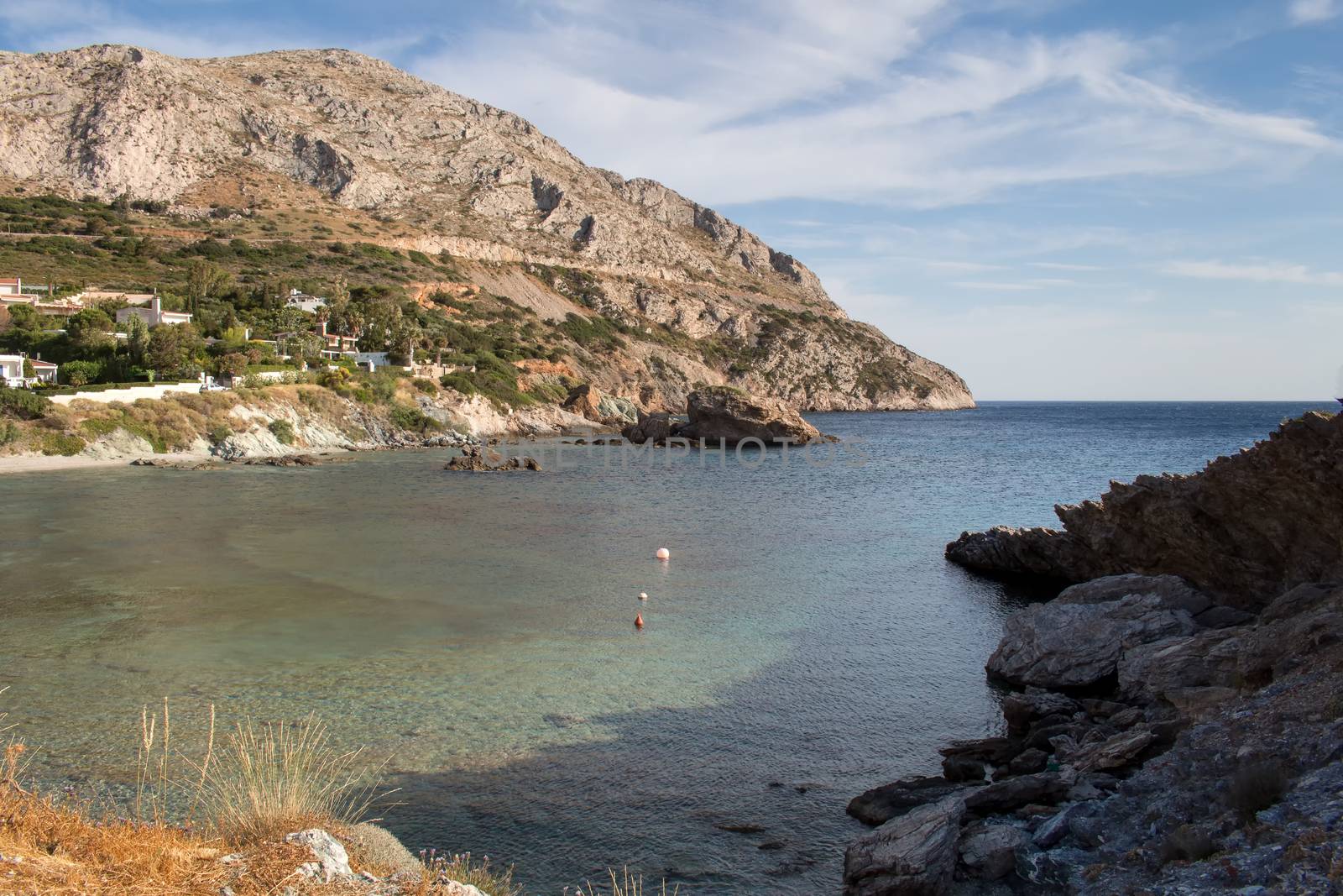 Peninsula of the greek coast. Rocky mount, partly covered with trees. Several houses. Sea bay. Clear water. Cloudy sky.