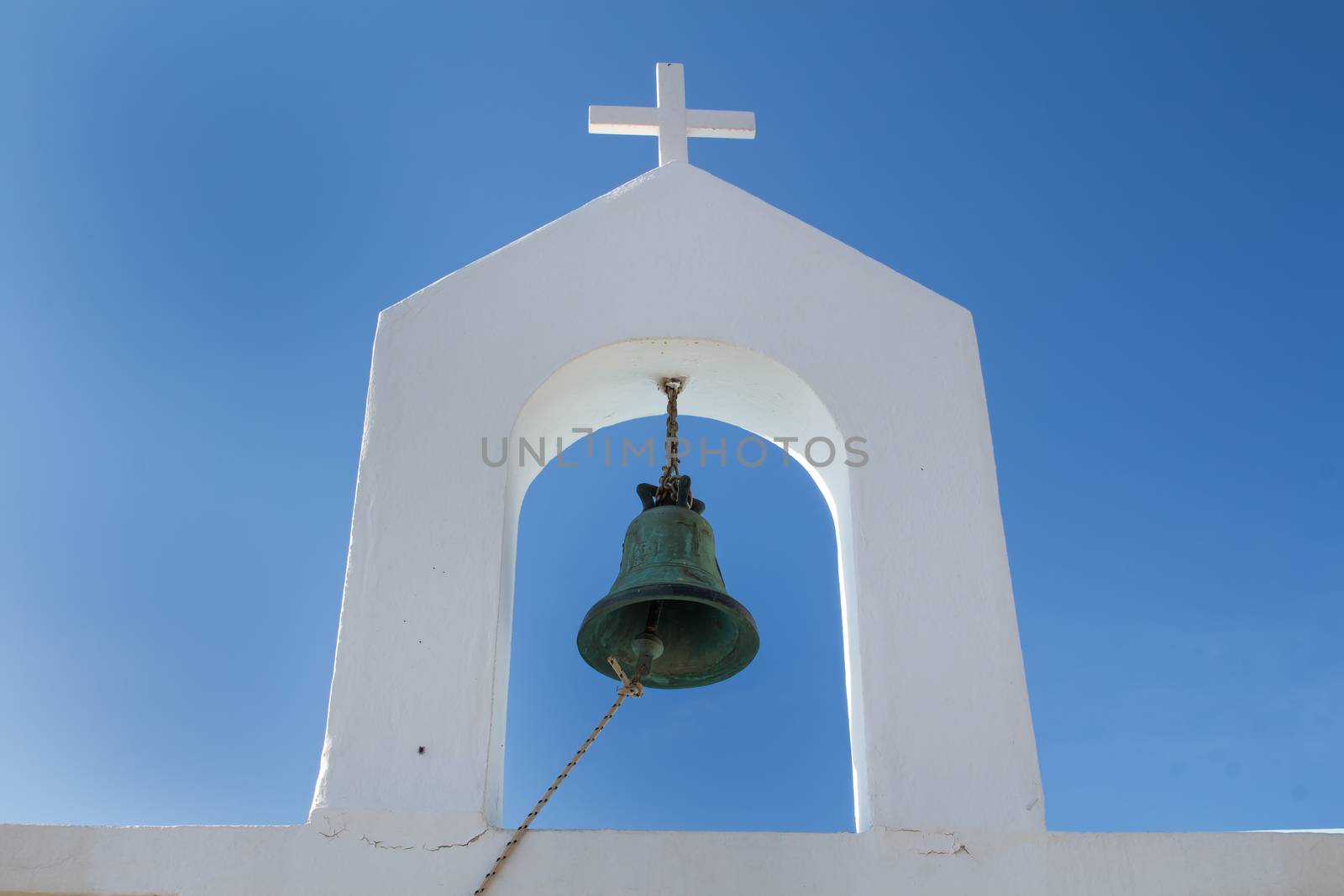 Detail of the architecture of a chapel in Greece. Heavy bell with a rope to use it. White tower with a cross. Bright blue sky in the background.