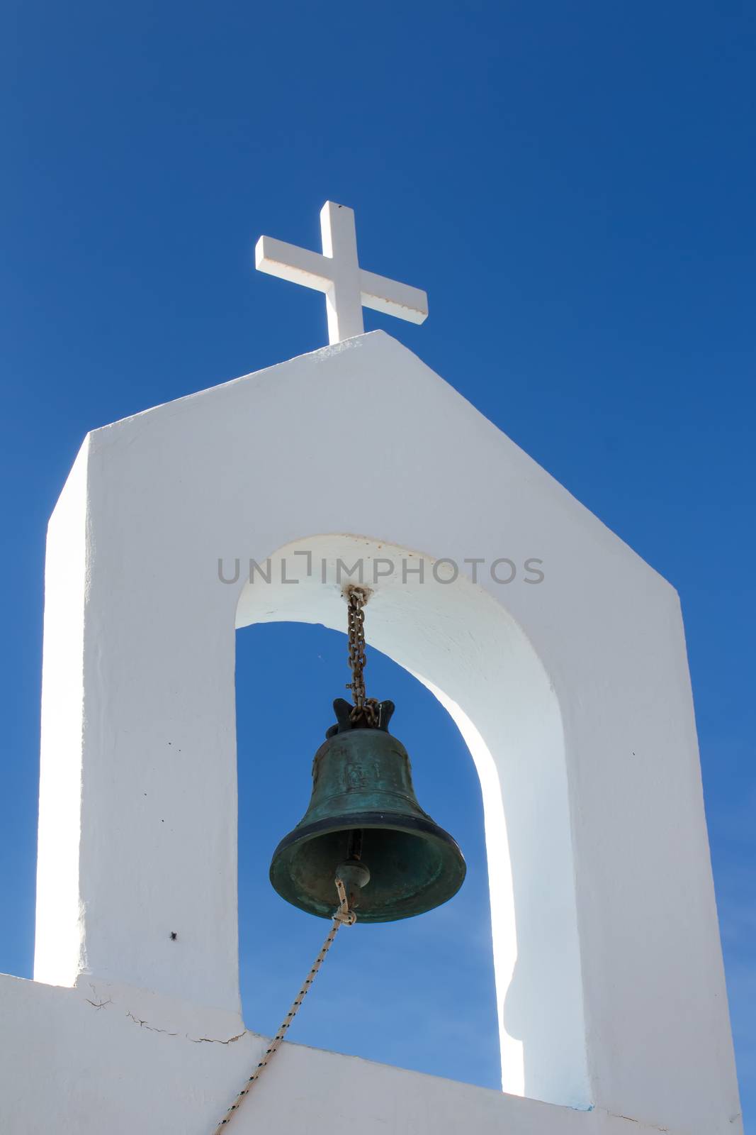 Detail of the architecture of a chapel in Greece. Heavy bell with a rope to use it. White tower with a cross. Bright blue sky in the background.