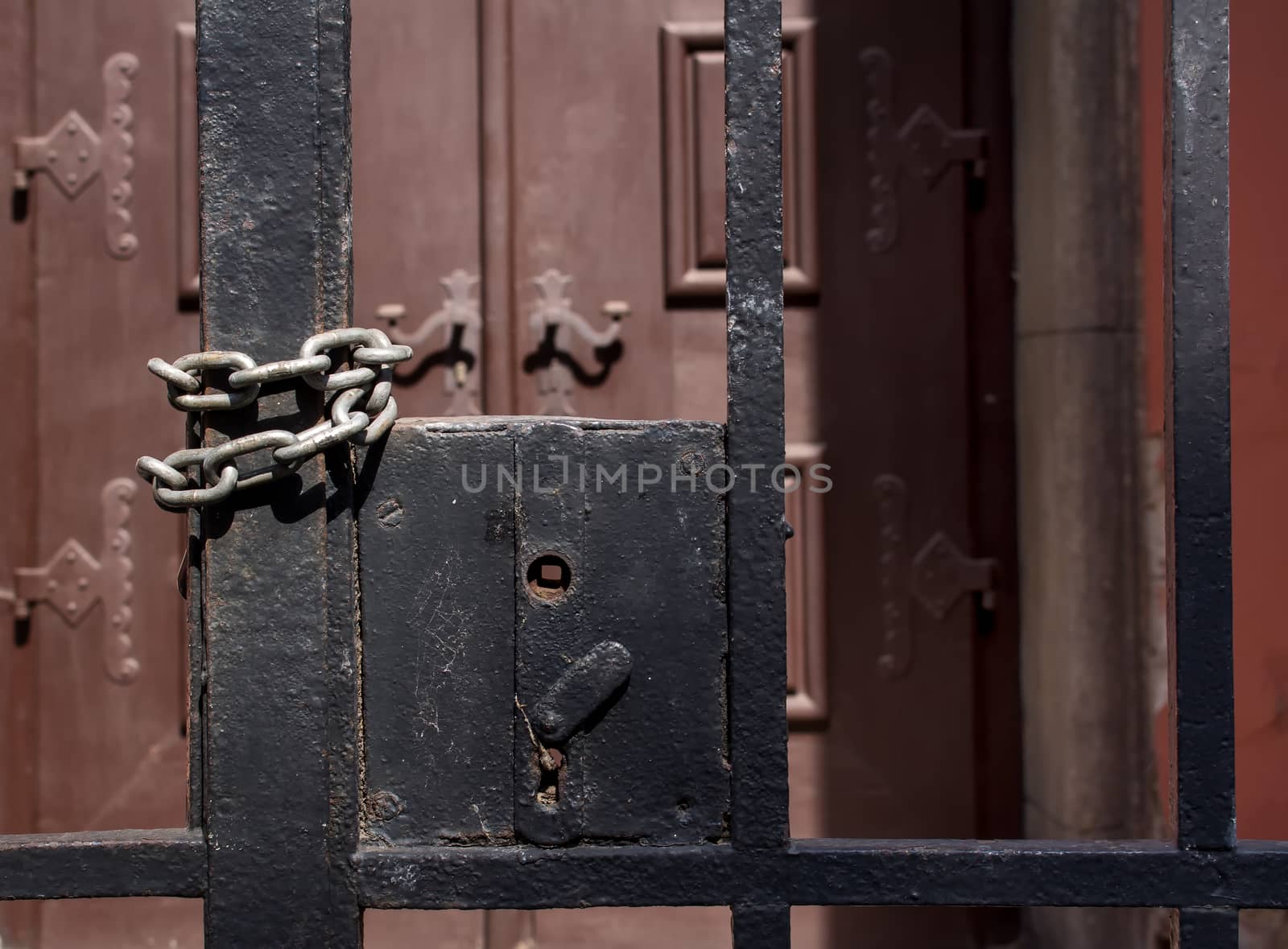 Grid fence with a missing handle. Twisted chain to keep it closed. Massive wooden decorated gate in the background.