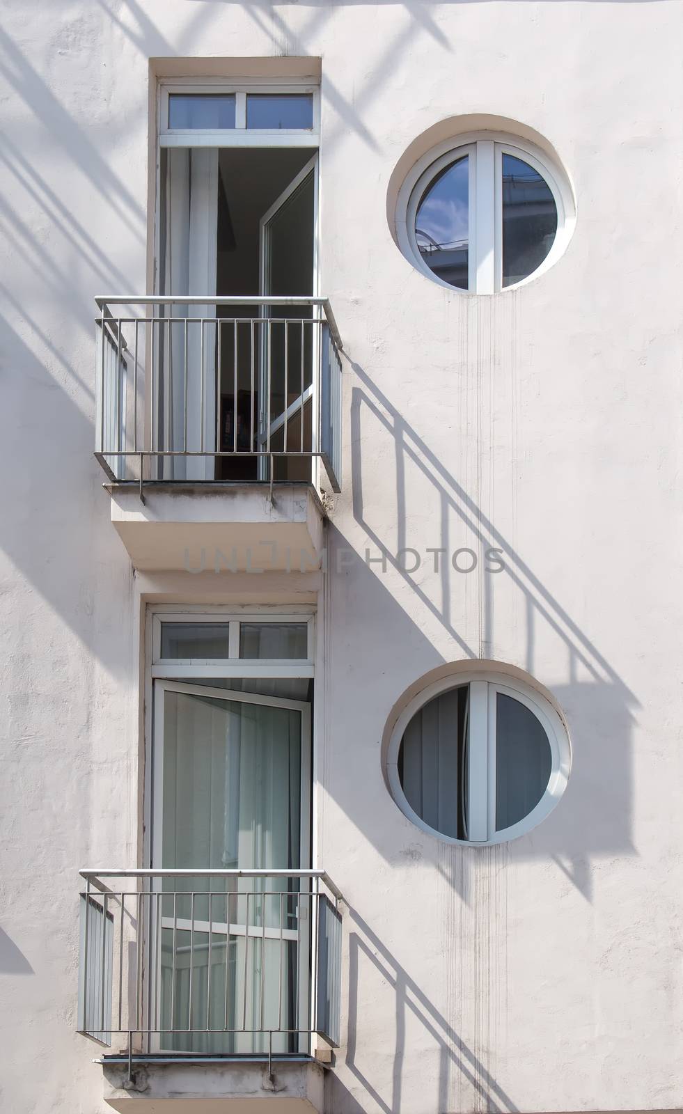 Detail of a white building with two balconies and circle windows. Lines of the shadows on the wall. Reflection of the sky in the windows.