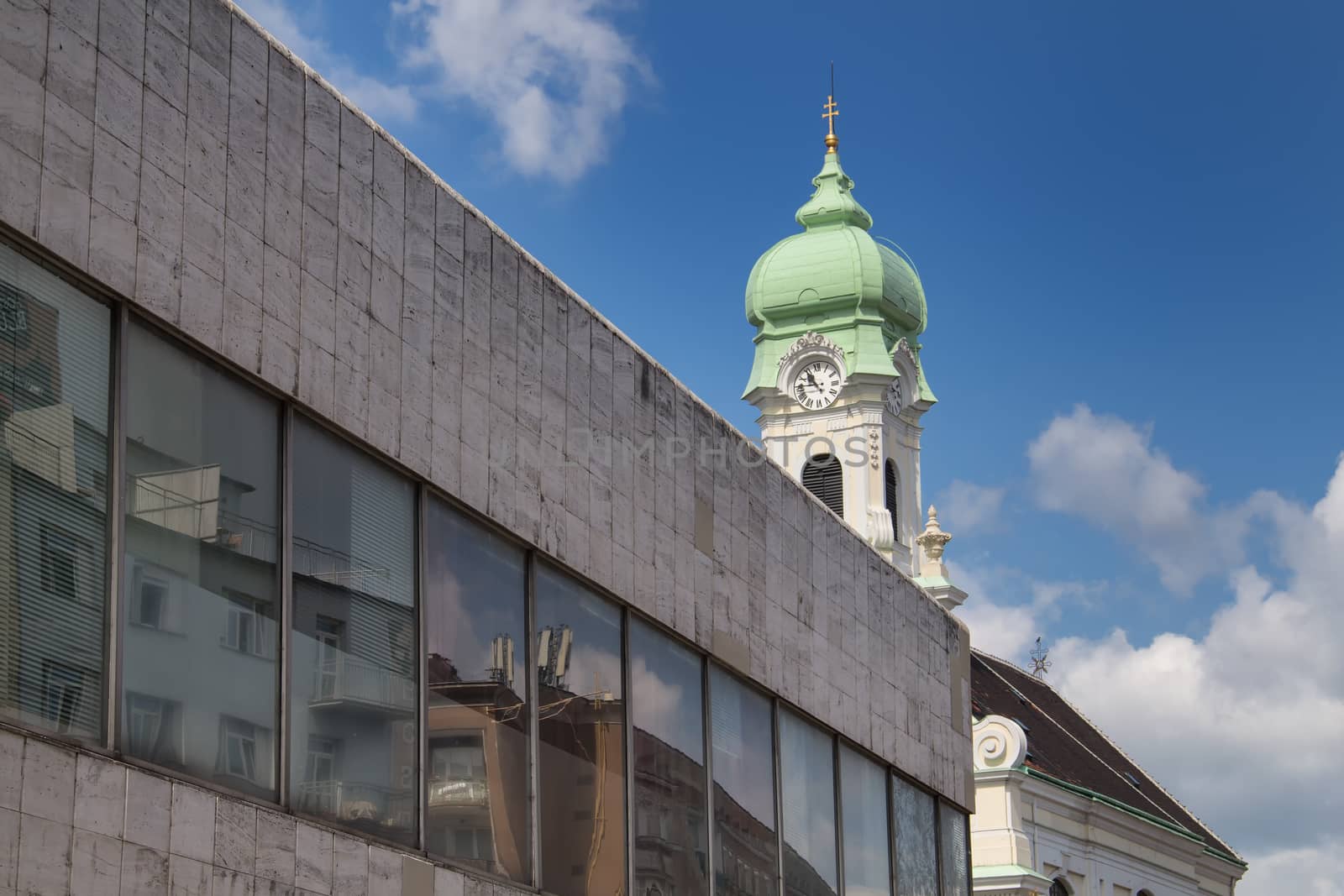 Contrast of a modern architecture made of marble and glass with a tower of baroque church in the background. Blue sky with white clouds.