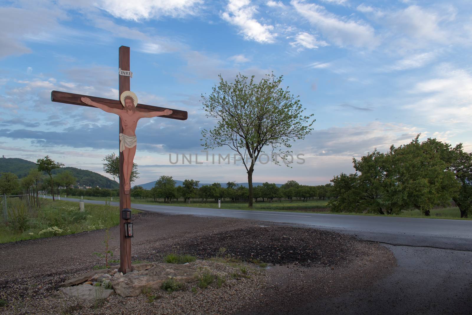 Early spring morning after a rain. Crucifix beside a road. Trees and fields in the background. Cloudy morning sky.