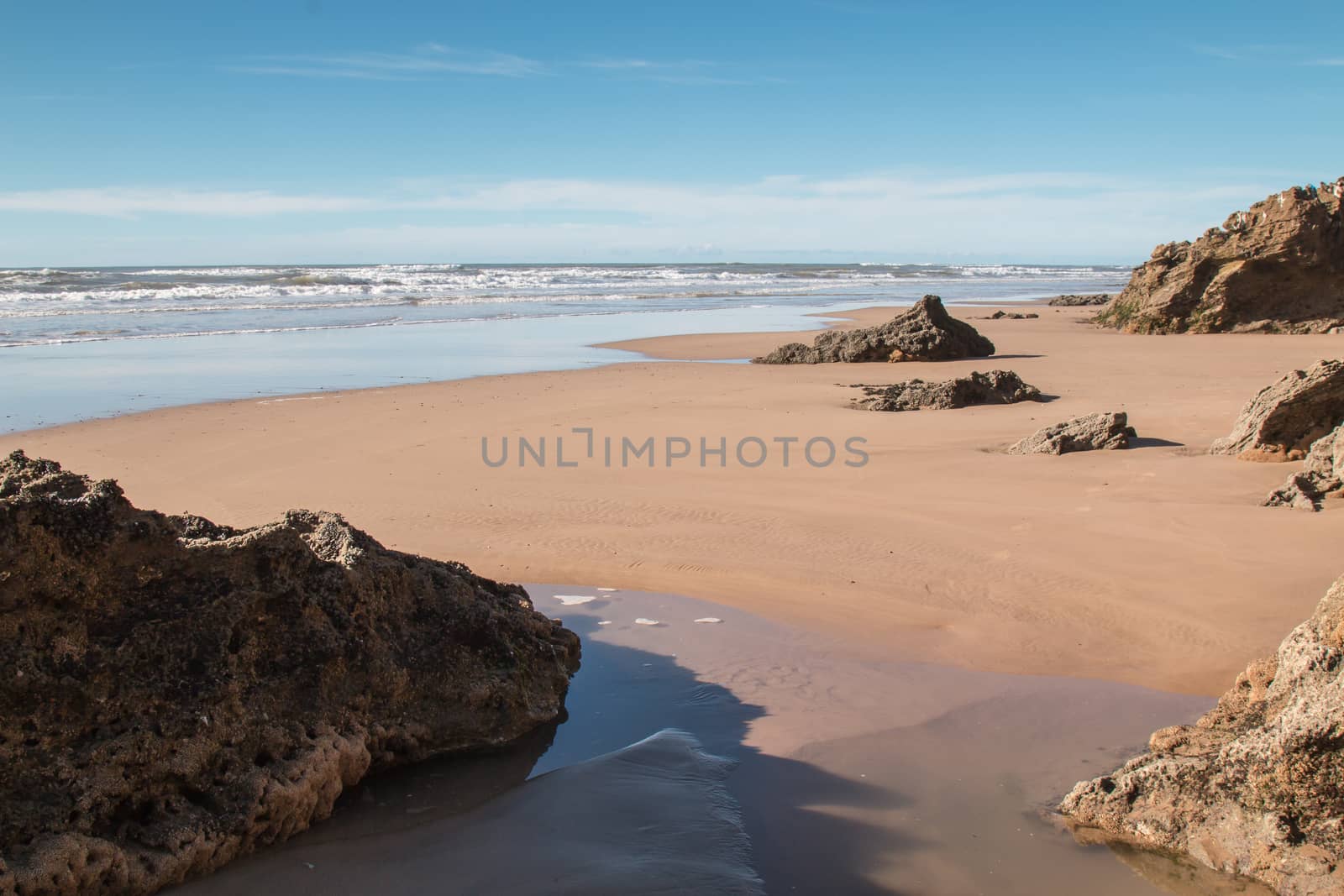 Sandy beach on the coast of Atlantic Ocean in Morocco. Wet sand, small waves, rocks in the foreground, bigger in the background. Cloudy sky.