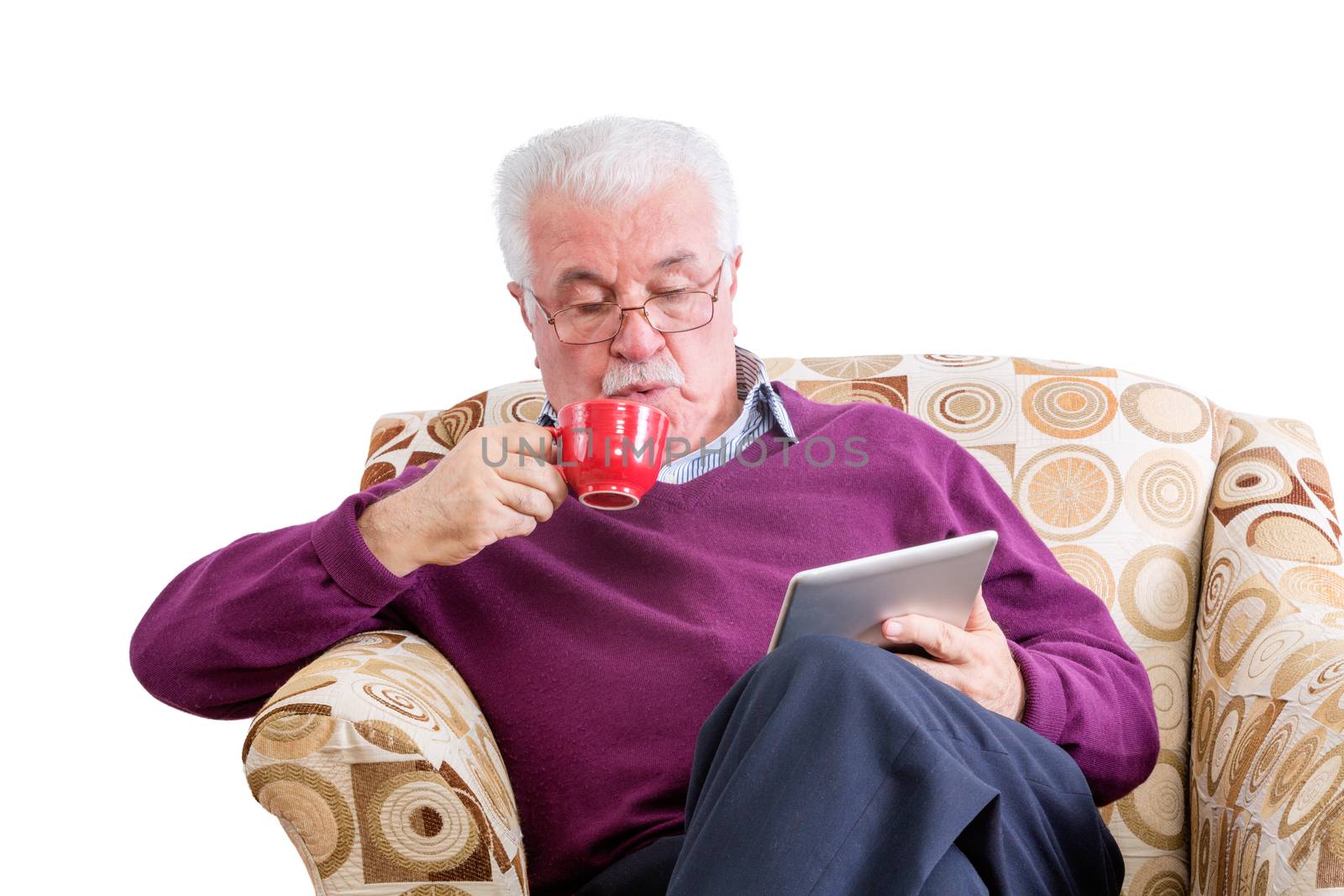 Senior male in purple sweater with mustache looking down at tablet computer while sipping from red mug and sitting in chair over white background