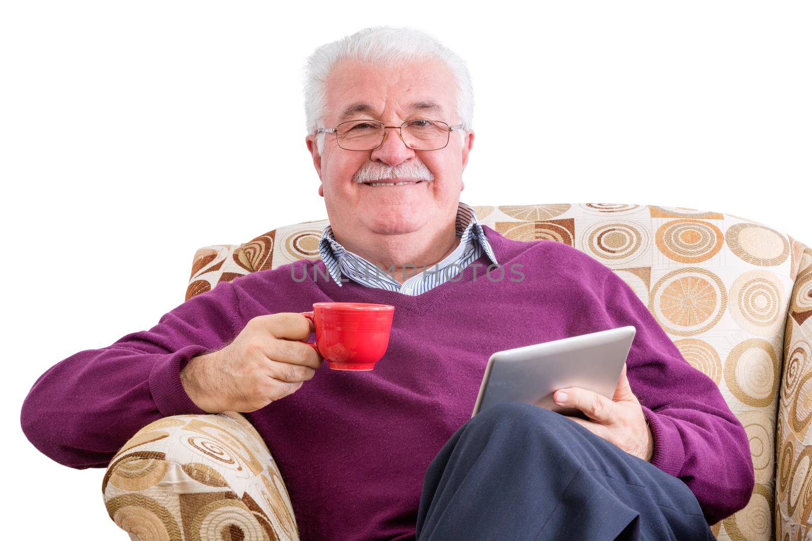 Cheerful senior male in purple sweater holding tablet computer in one hand and a red mug in the other while relaxing in chair over white background