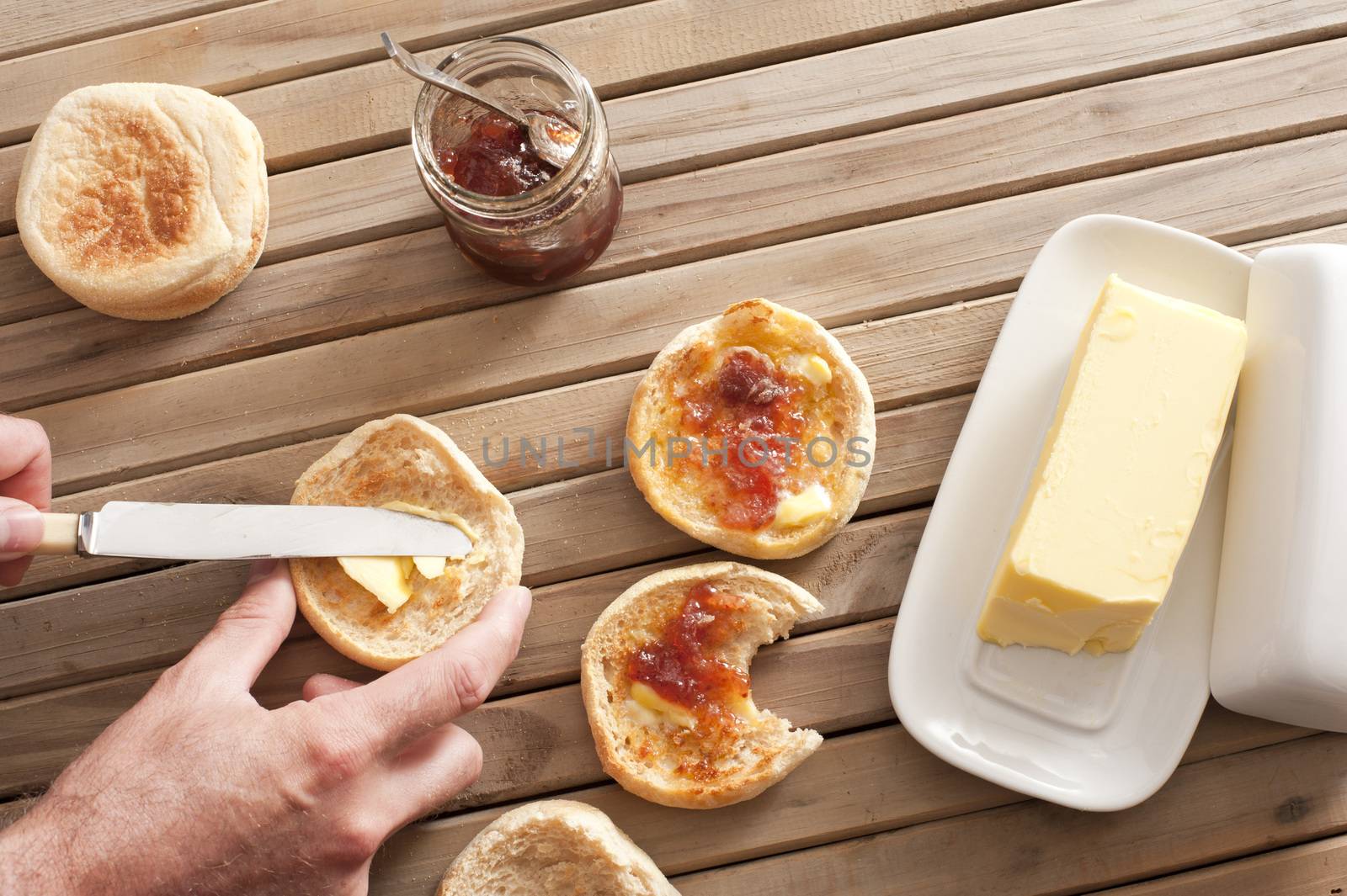 Man buttering a freshly baked toasted crumpet for breakfast from a large pat of butter with a jar of preserve or jam alongside, overhead view of his hands