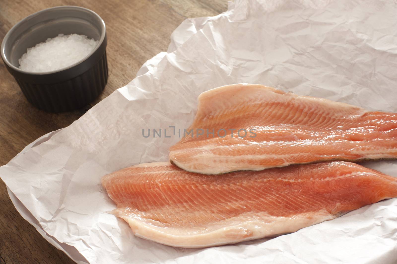 Two fresh raw rainbow trout fillets on crumpled white paper for a delicious seafood dinner with a small dish of coarse salt alongside