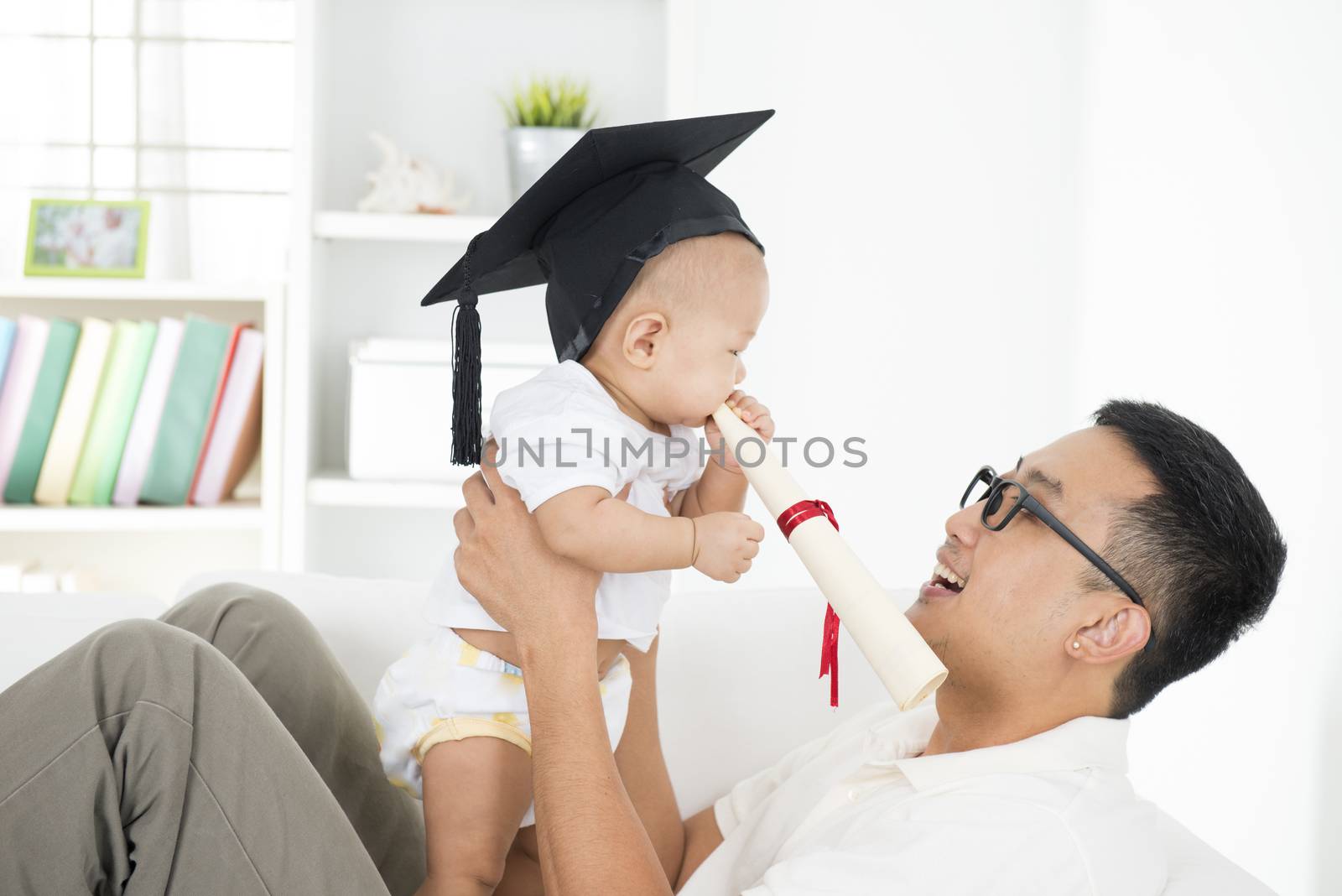 Baby with graduation cap holding certificate with father. Parent and child early education concept. Asian family lifestyle at home.