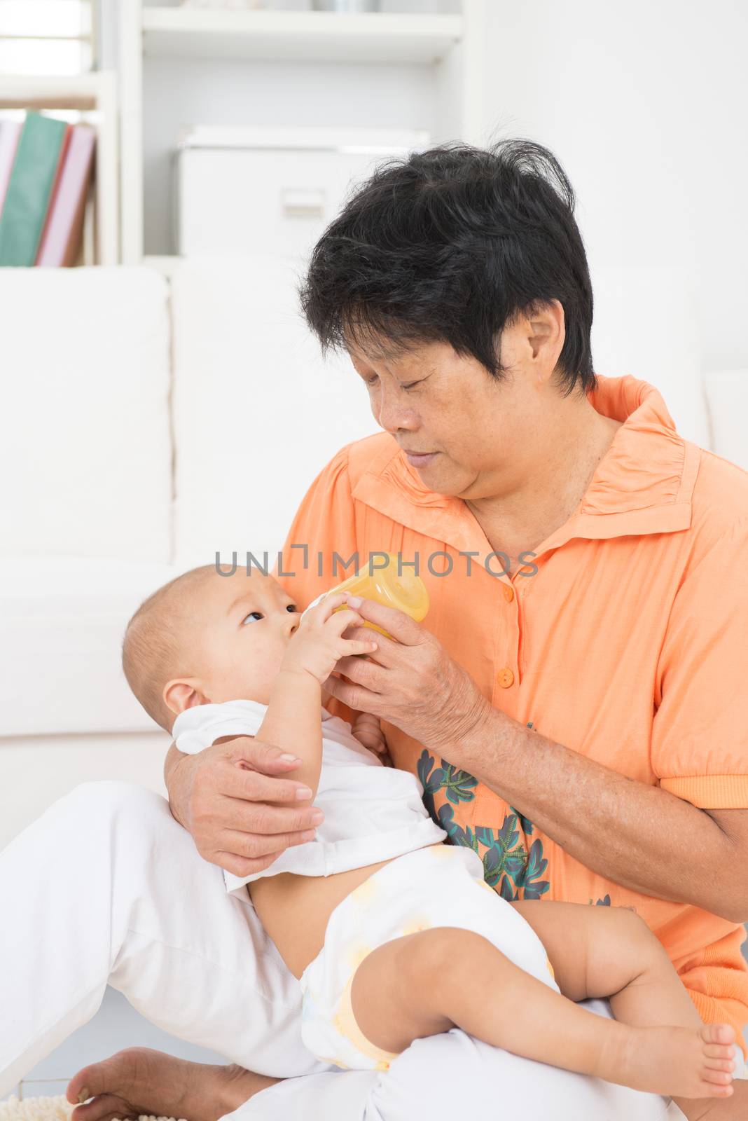 Grandmother taking care grandchild at home. Feeding solid food fruit puree with bottle.