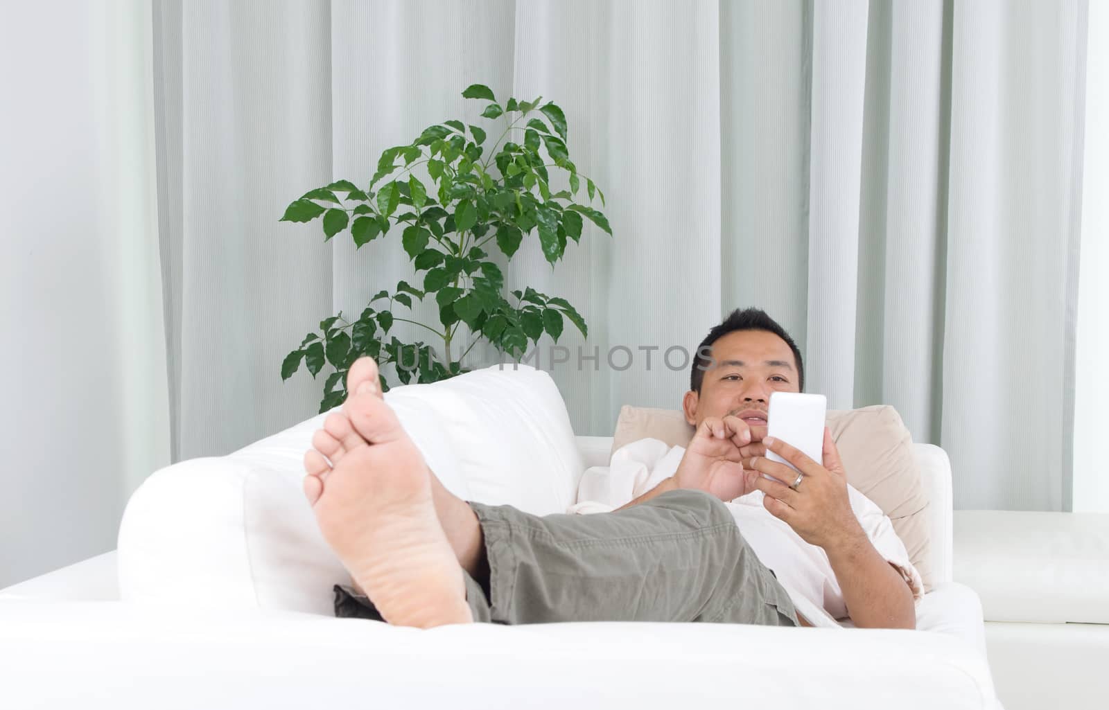 Smartphone mobile apps concept. Malay guy using smart phone. Handsome asian man relaxed and lying on sofa indoor. 