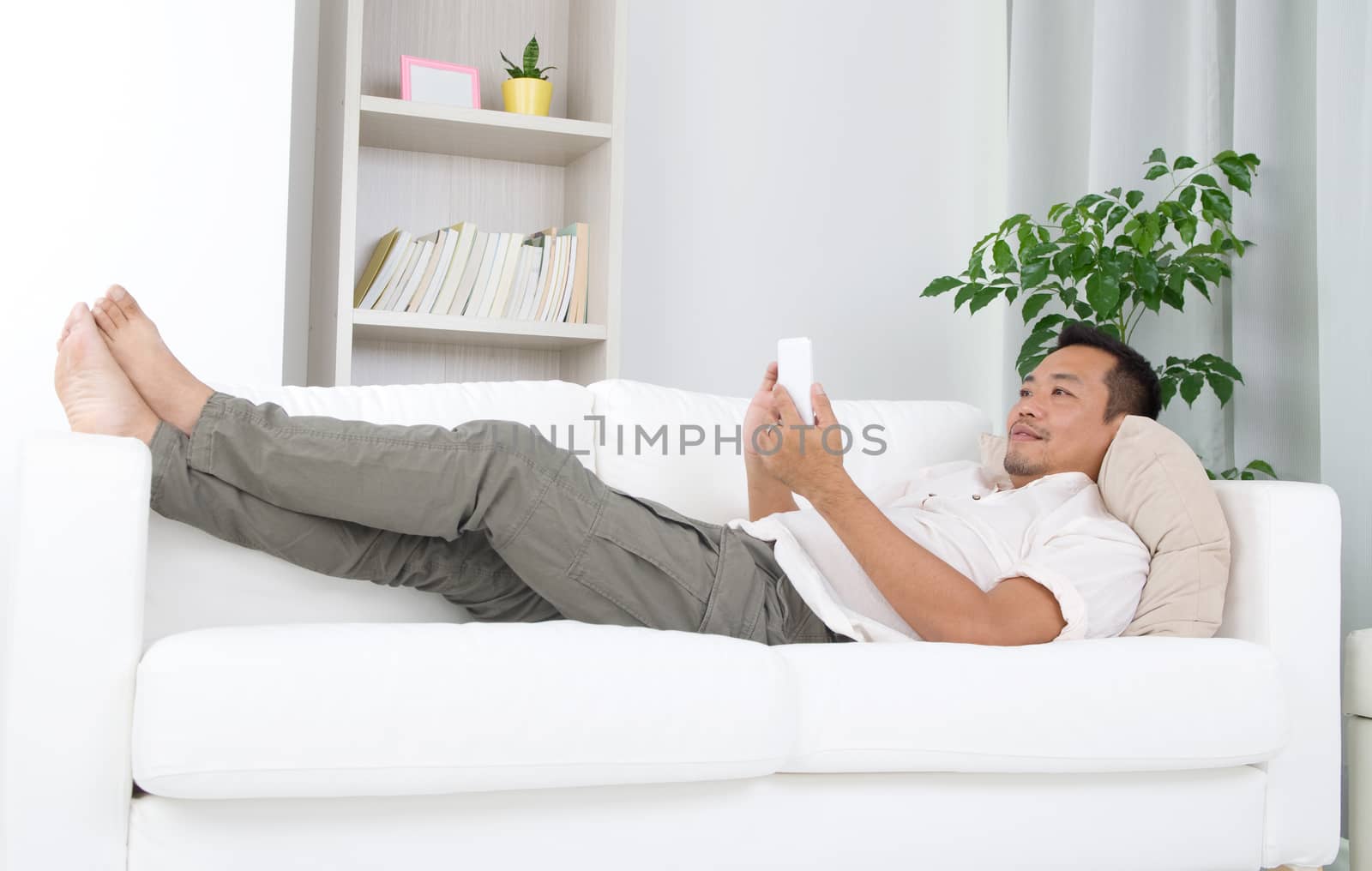 Smartphone mobile apps concept. Malay guy using smart phone. Handsome asian man relaxed and lying on sofa indoor. 