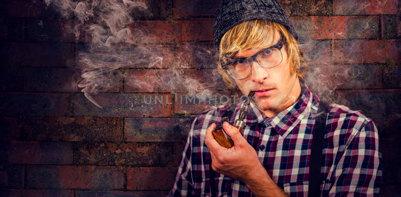 Serious blond hipster smoking a pipe against texture of bricks wall