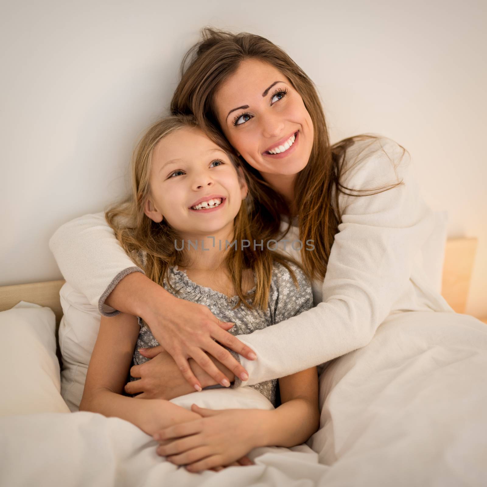 Beautiful smiling mother and her daughter hugging in bed. They are sitting on bed at pajamas and smiling.