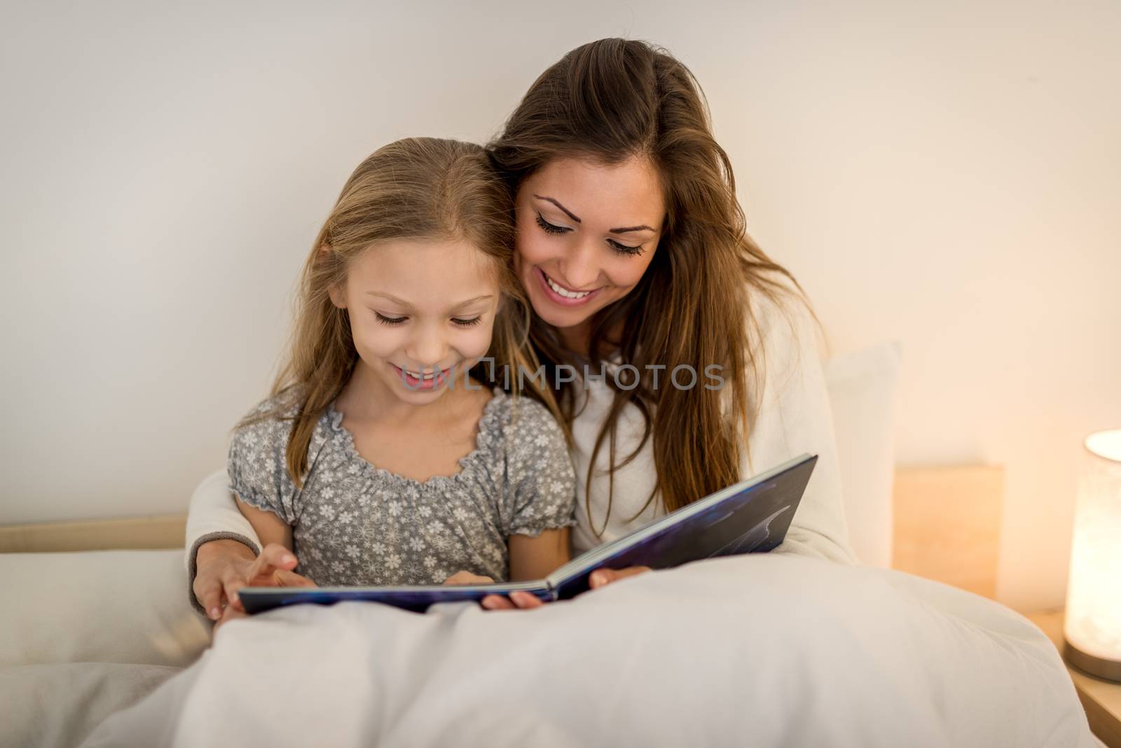 Beautiful smiling mother and her daughter reading book in bed. 