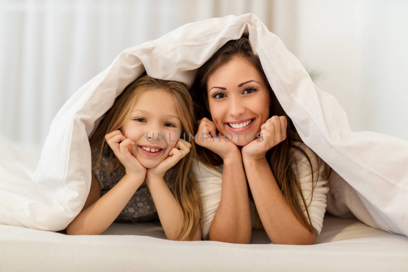 Beautiful smiling mother and her daughter having fun in bed. They lie in bed looking at camera and peeking under the blanket with smile.