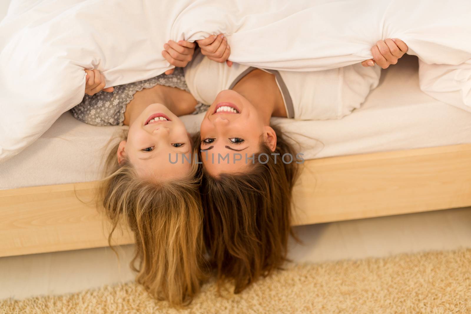 Beautiful smiling mother and her daughter having fun in bed. They lie in bed and looking at camera with smile.
